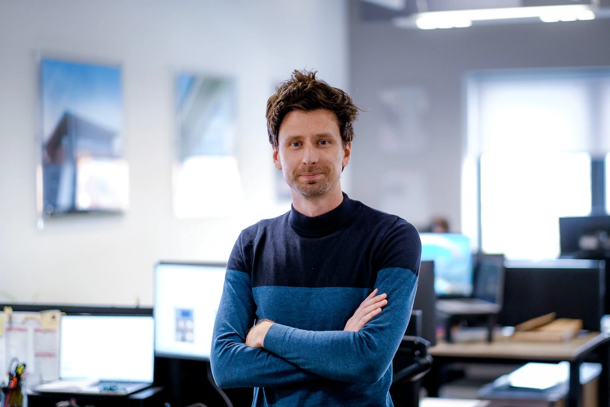 A man with dark hair and a slight beard wearing a brown jacket and gray sweater standing in an office with work desks and other employees in the background.