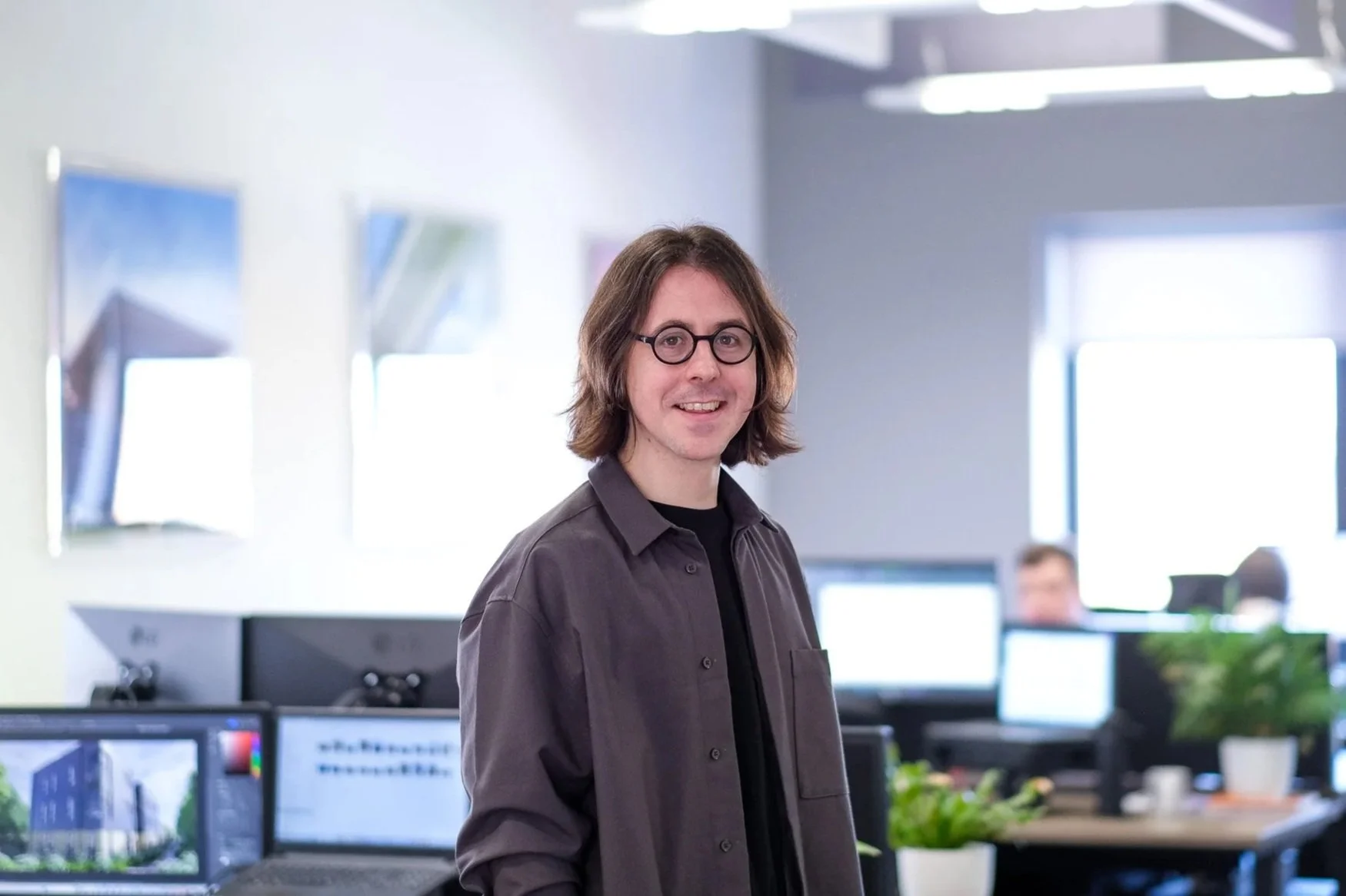 A man with glasses and shoulder-length brown hair, smiling in a modern office with computers, desks, and potted plants.
