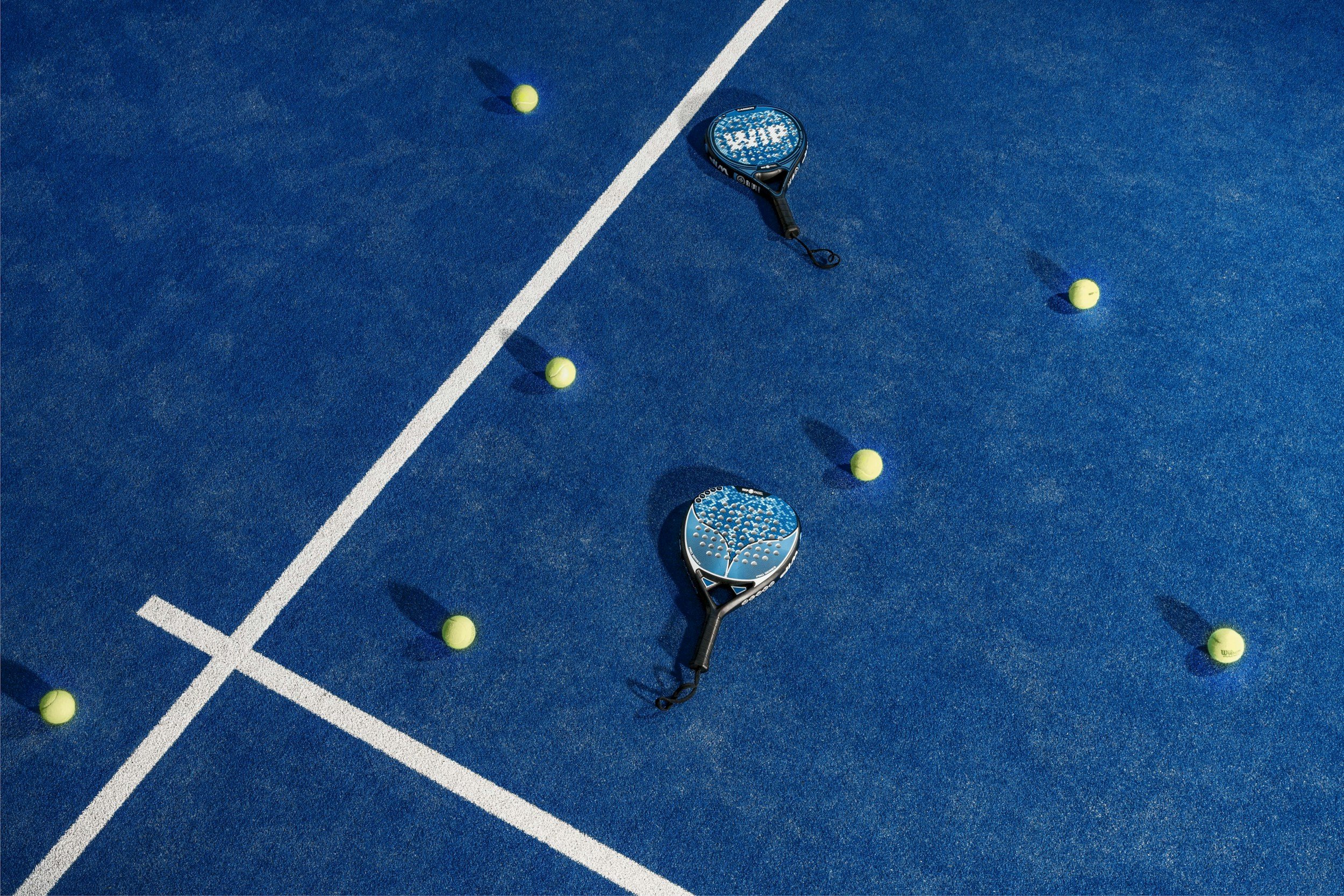 Deux raquettes de padel et plusieurs balles de tennis jaunes dispersées sur un court de tennis en bleu avec des lignes blanches.