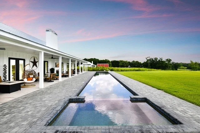 A modern house with a long, narrow in-ground pool in the backyard, surrounded by a paved patio area with outdoor furniture, green lawn, and a colorful sky at sunset.