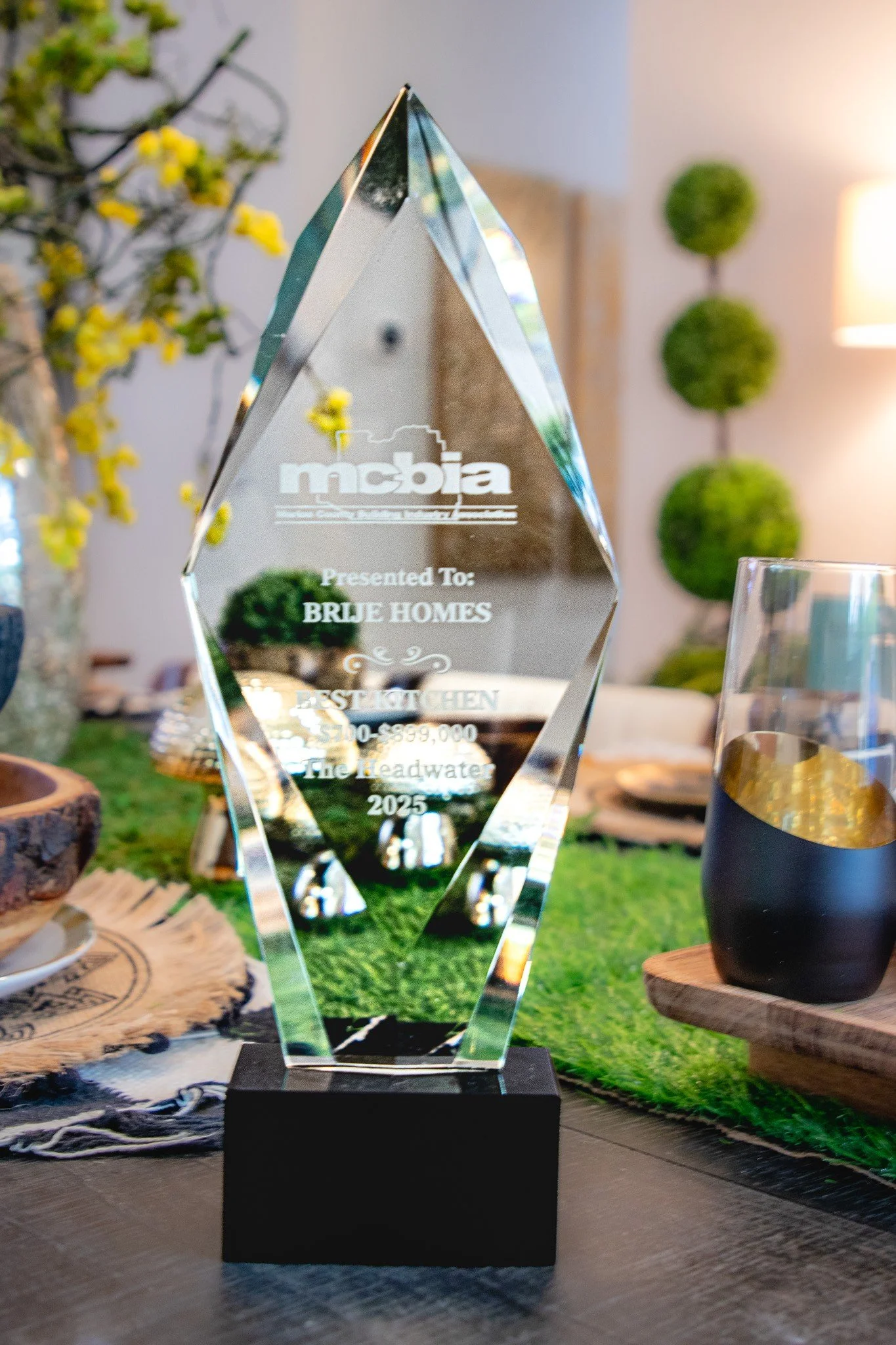 A crystal award with engraved text sitting on a black base, displayed on a table with various decorative items and greenery in the background.