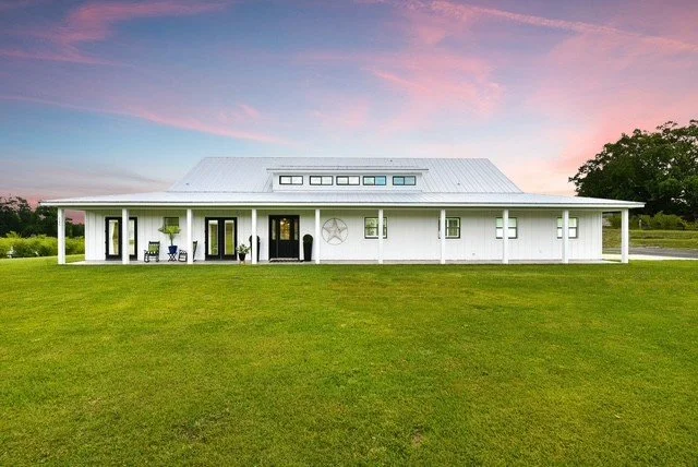 White house with a porch, located in a grassy area during sunset with pink and blue sky.