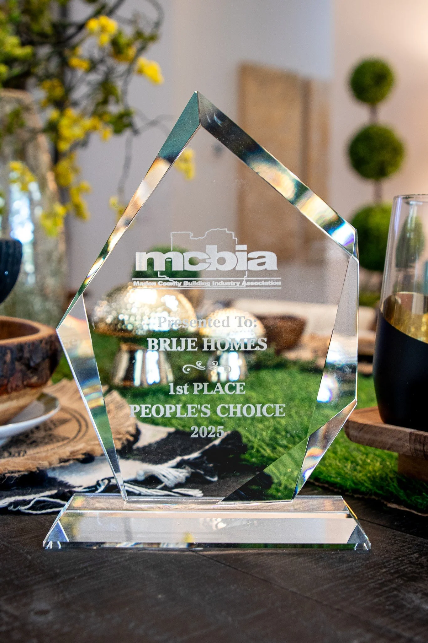 A crystal award with the logo of the Marion County Building Industry Association, awarded to Brije Homes for 1st place People's Choice in 2025. The award is on a wooden table with decorative objects and greenery in the background.