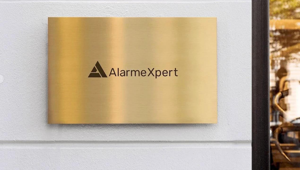 Plaque en métal doré avec le logo et le nom 'AlarmeXpert' affichés sur un mur blanc.