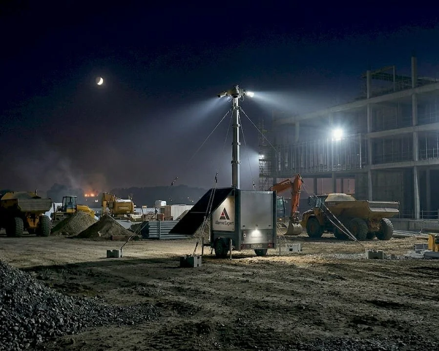 Chantier de construction la nuit avec éclairage, excavatrice, camions, et bâtiment en construction.