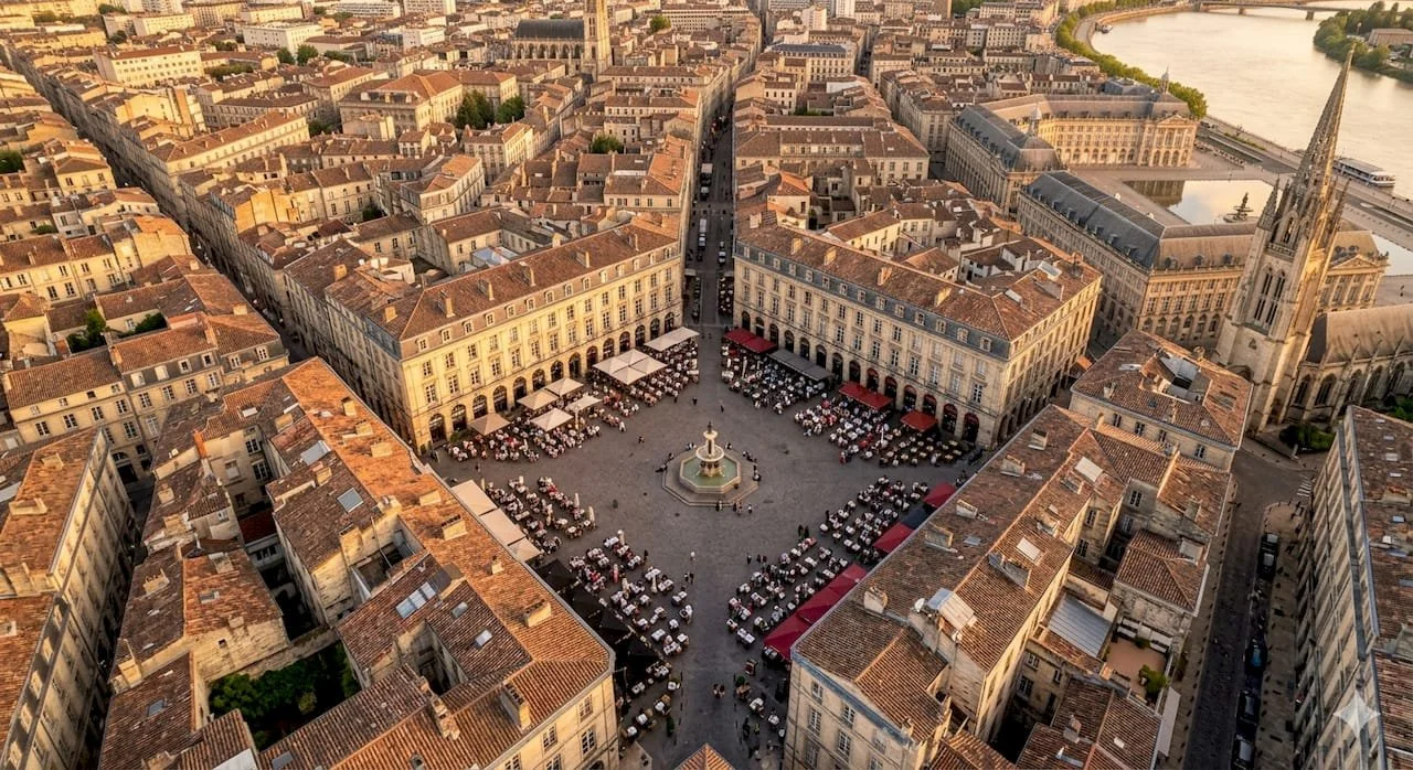 Vue aérienne d'une place urbaine avec une fontaine au centre, entourée de bâtiments historiques et de cafés avec tables. La place est située dans une ville européenne près d'une rivière.