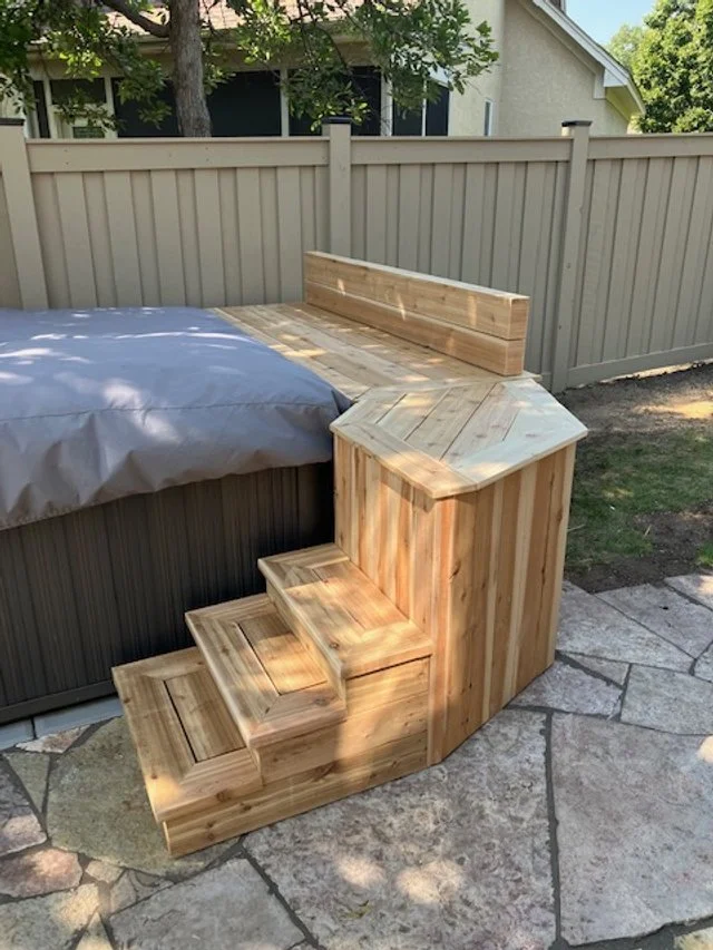 A wooden built-in step and bench beside a hot tub outdoors, with a vinyl fence and trees in the background.