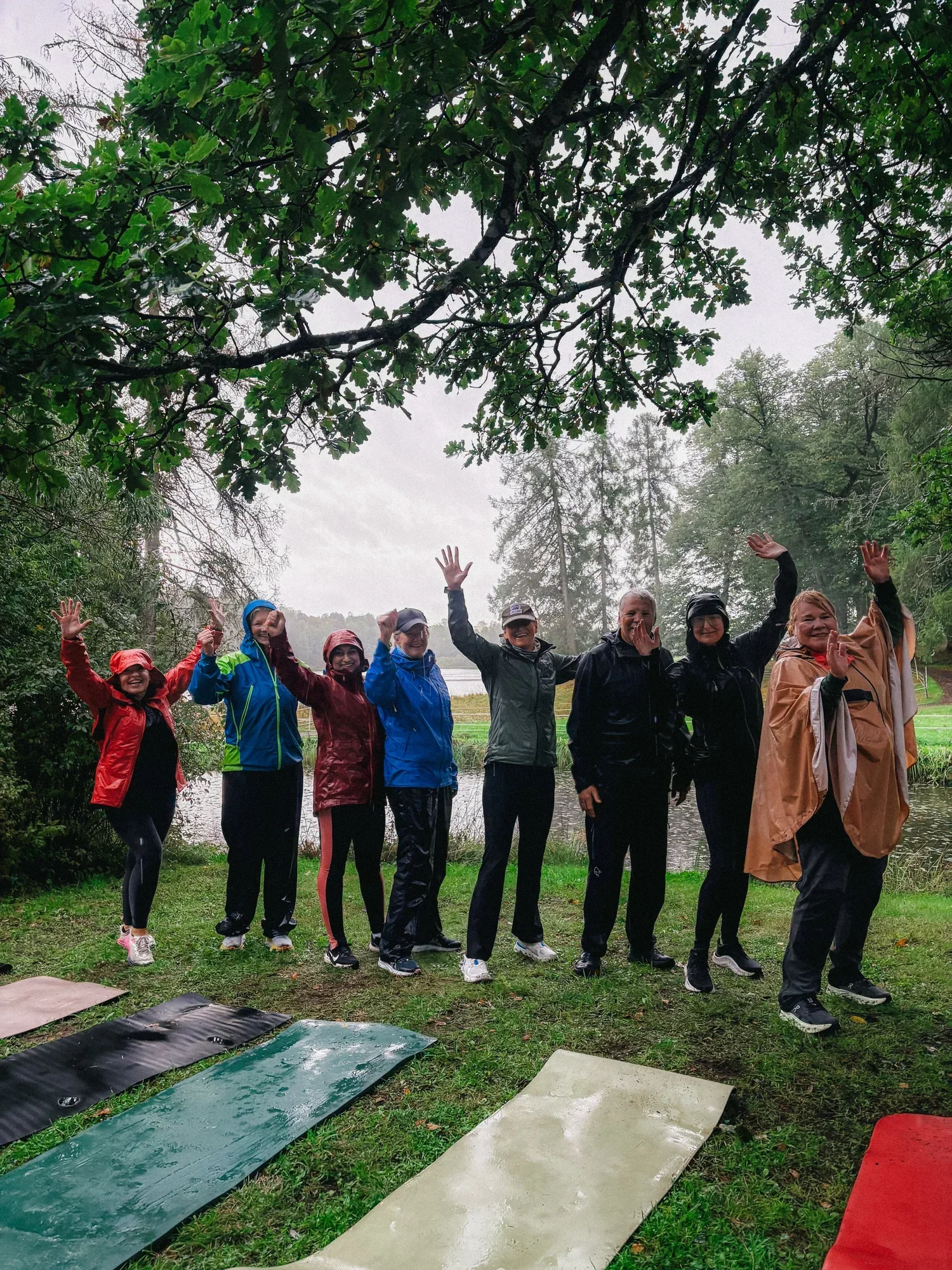 En gruppe mennesker som har på seg regnfrakker og er ute i regnet under trærne, og de vinker til kameraet.