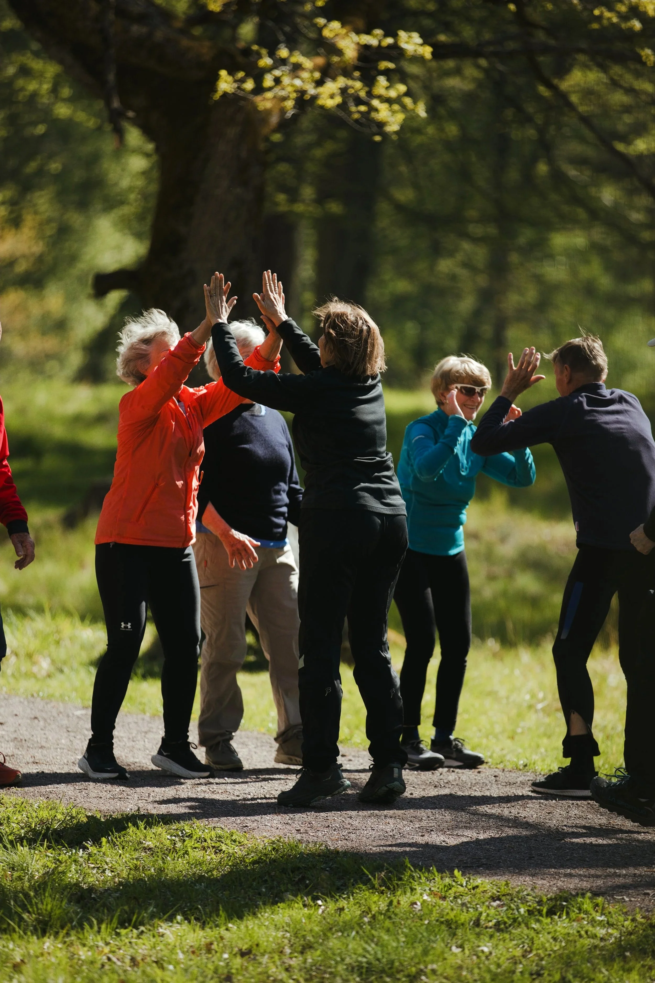 En gruppe eldre mennesker trener utendørs på en grussyte, hvor de ligger på yogamatte og gjør øvelser ved bruk av manualer. Det er høst med høstfargede blader på bakken og trær i bakgrunnen.