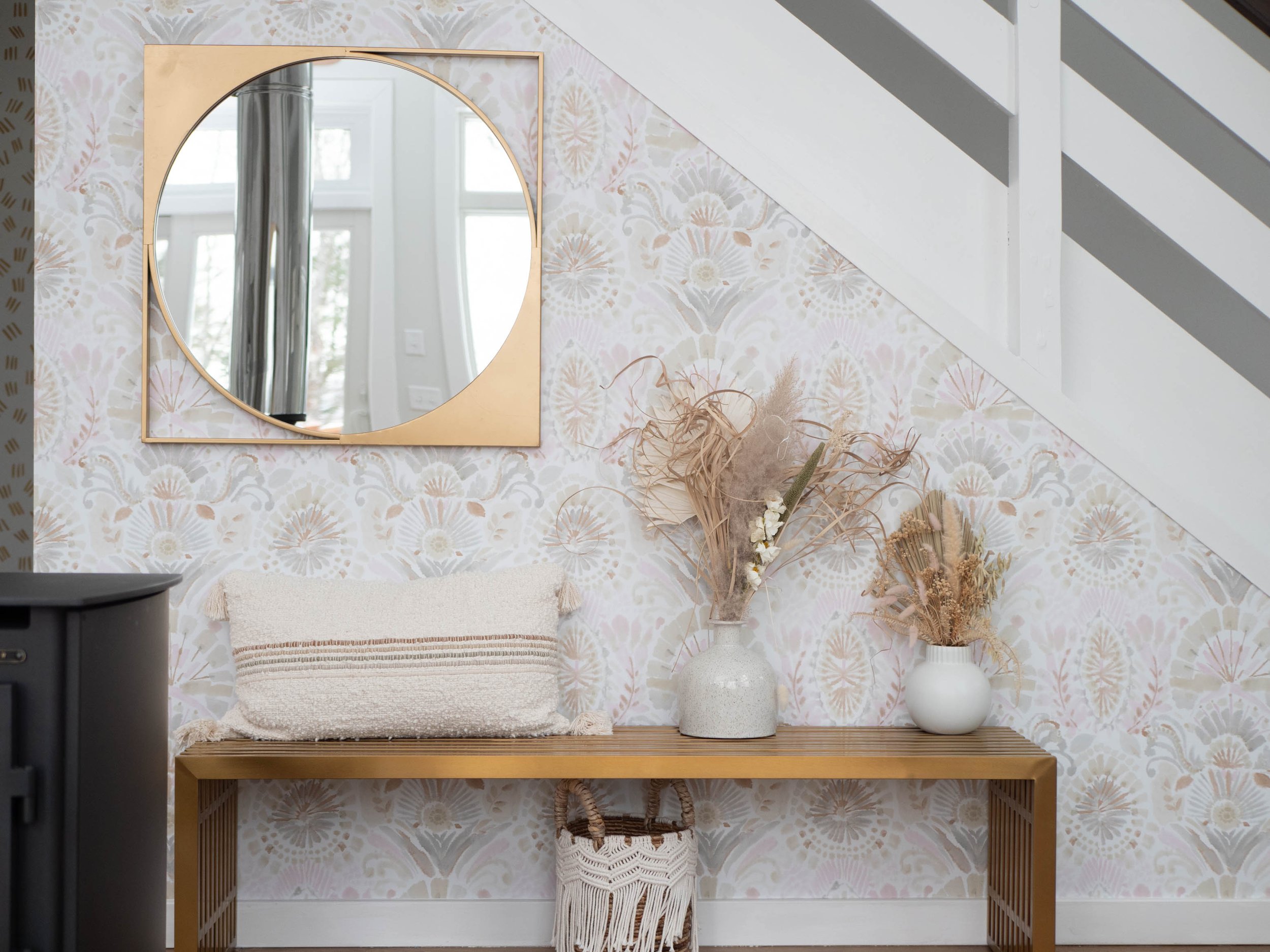 Decorative wall under staircase with a square and round mirror, wooden console table, and two white vases with dried flowers.
