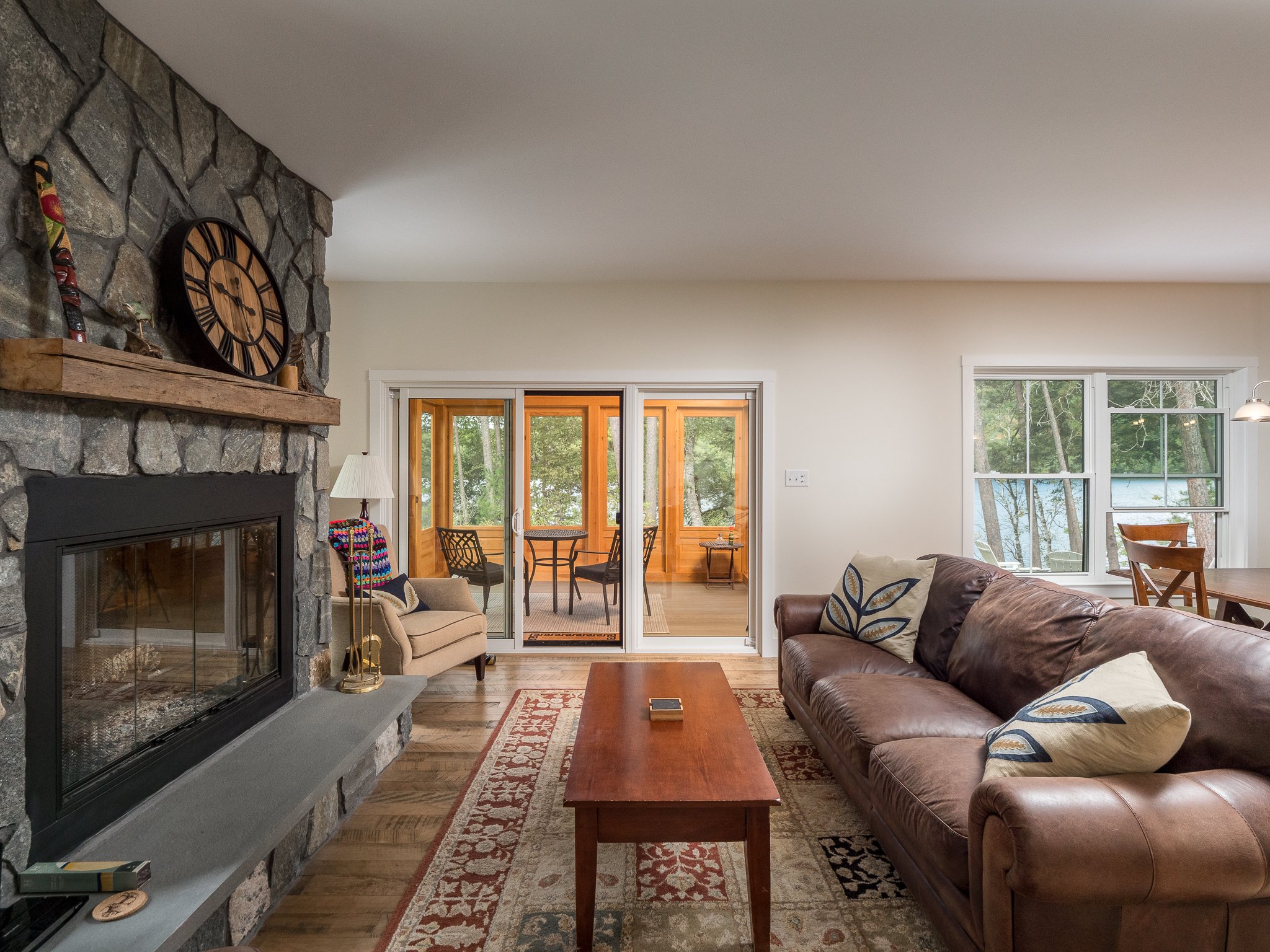 Cozy living room with stone fireplace, leather sofa, and large windows overlooking a lake. A wooden coffee table is centered on a patterned rug. A clock and decorative items adorn the mantel. Sliding doors open to a sunroom with a table and chairs.