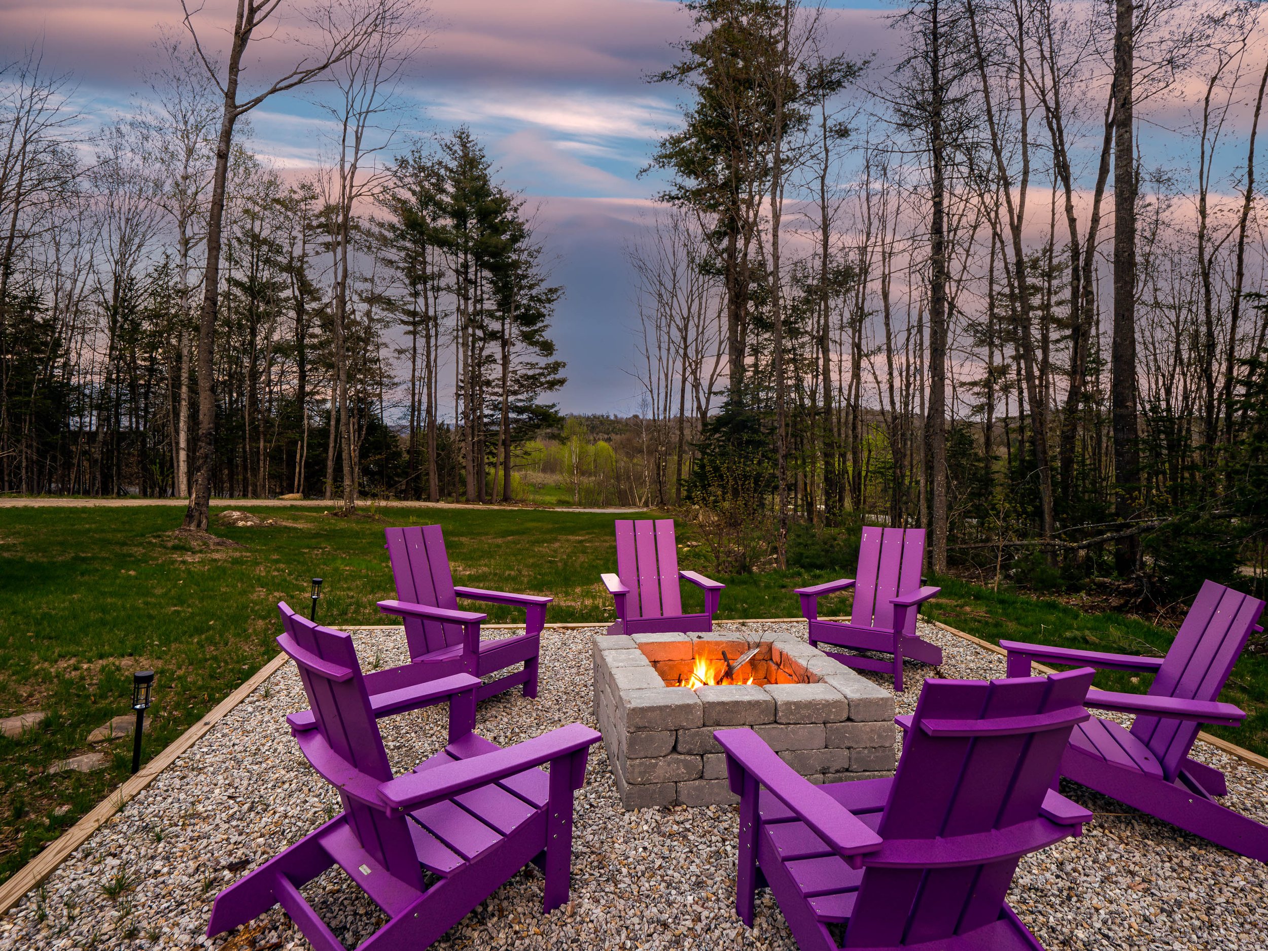 Six purple Adirondack chairs arranged in a circle around a small brick fire pit on a gravel patio, with a background of trees and a cloudy evening sky.