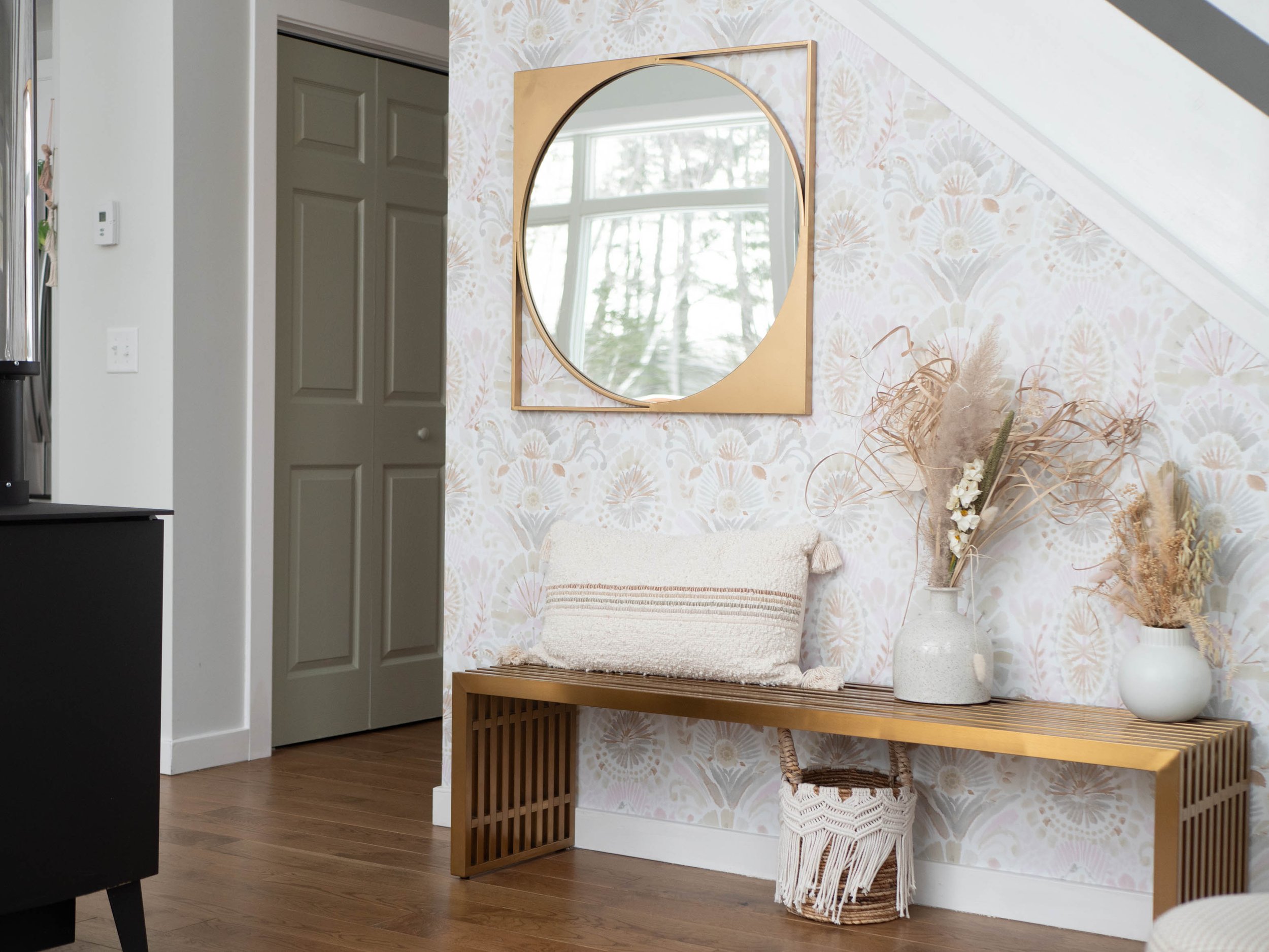 Interior of a room featuring a gold-framed mirror, a wooden bench with a decorative pillow, and vases with dried flowers on a floral wallpapered wall.