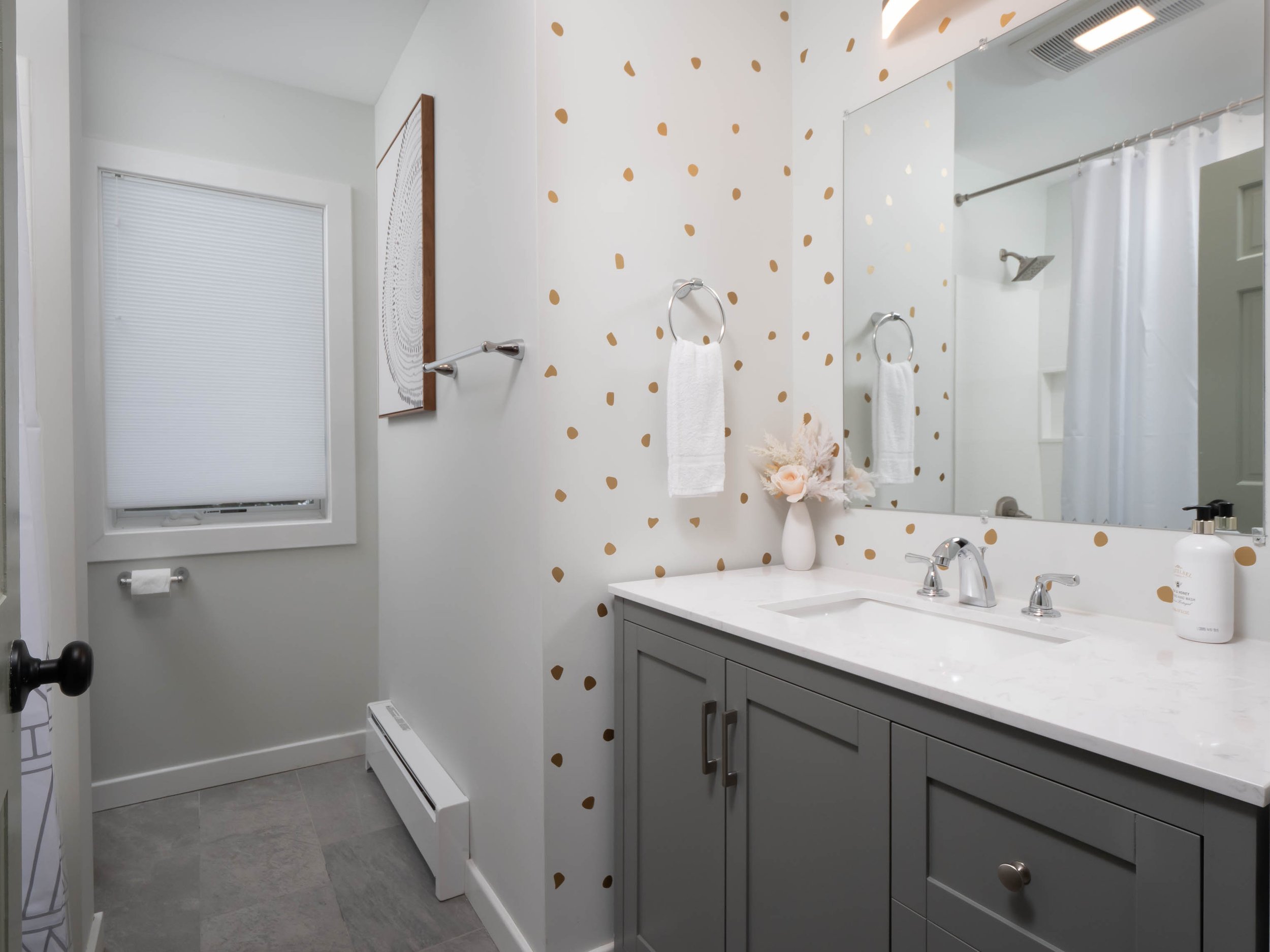Bathroom with gray vanity, white countertop, large mirror, white walls, gold dot wallpaper, window with white blind, white towel on ring, floral arrangement in white vase, shower with white curtain, gray floor tiles.
