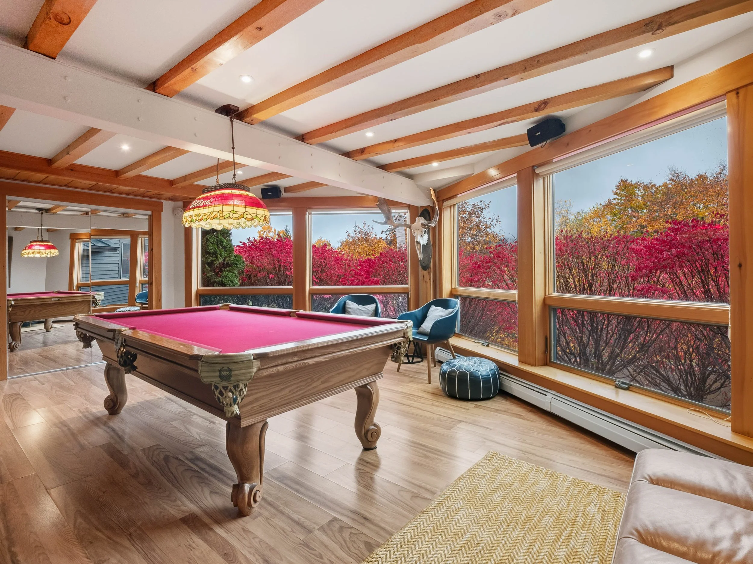 A cozy game room with a red pool table, large windows showing colorful autumn trees outside, wooden beams on the ceiling, and comfortable chairs.