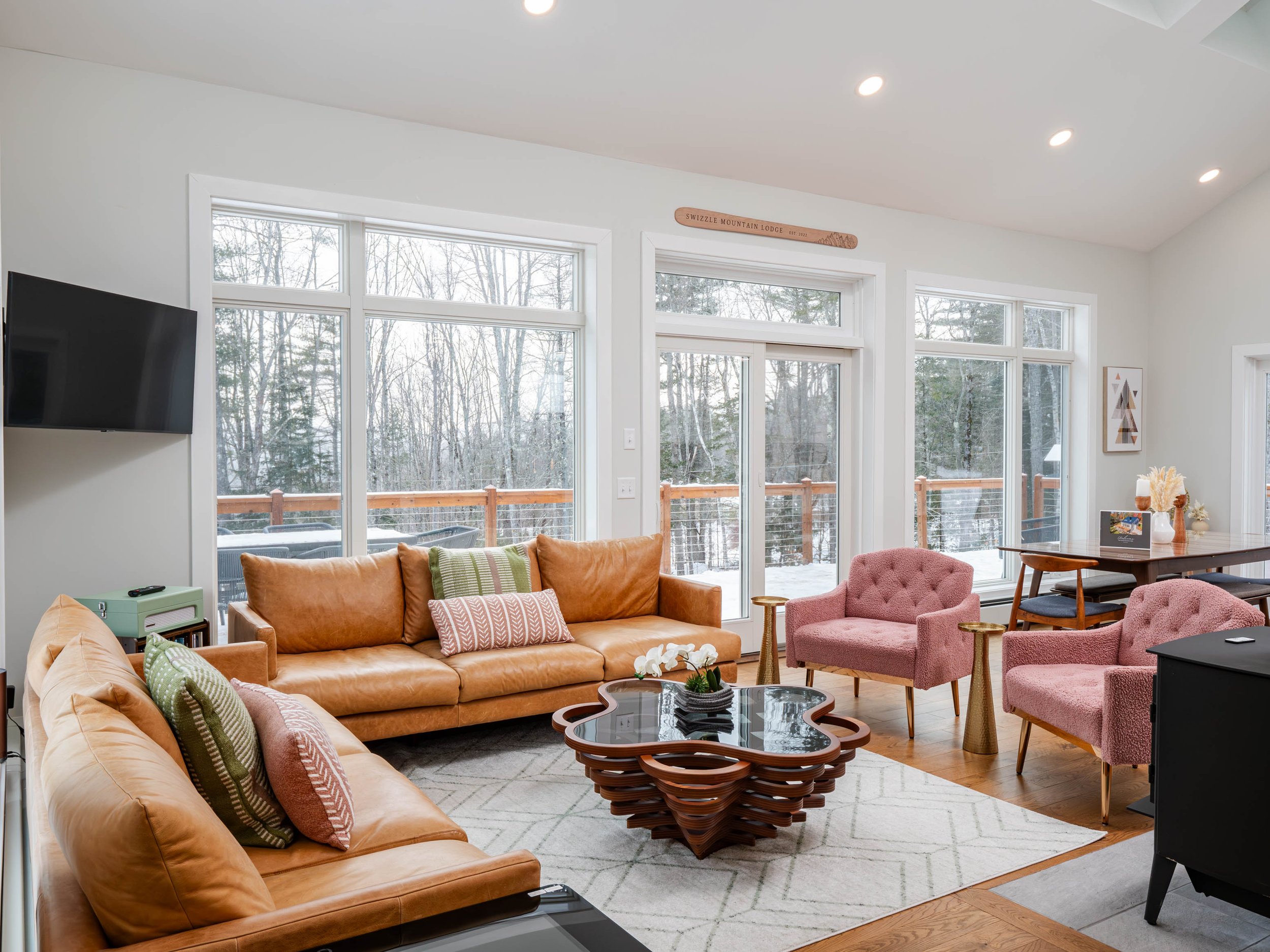 Living room with large windows showing snow outside, tan leather sofas, pink armchairs, a wooden coffee table, and a dining table in the background.