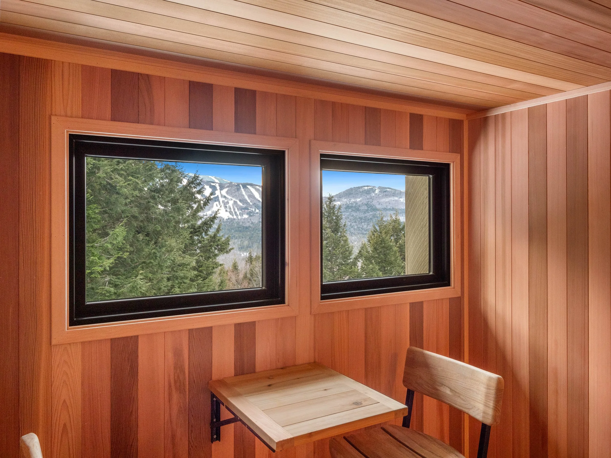 View of snowy mountain ski slopes and green trees through two side-by-side windows in a wooden interior room.
