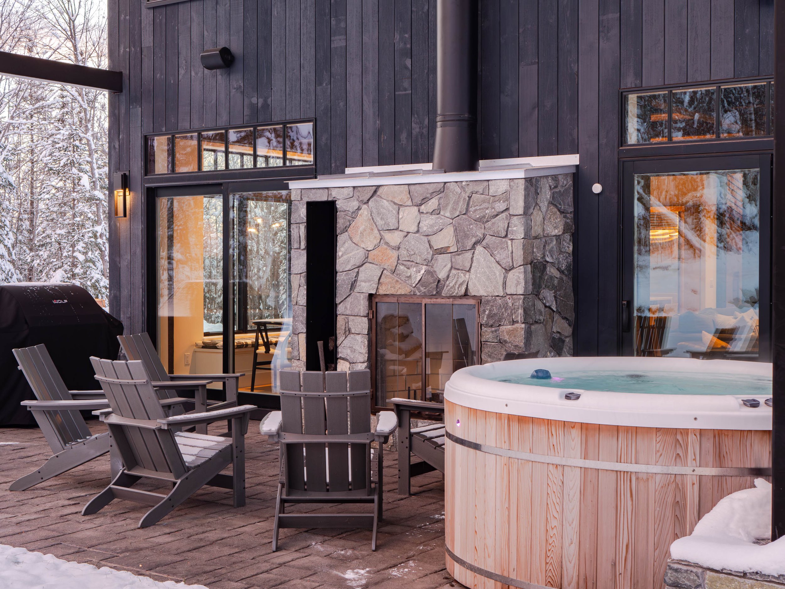 Outdoor patio with hot tub and chairs, stone fireplace, and snow-covered trees in the background.