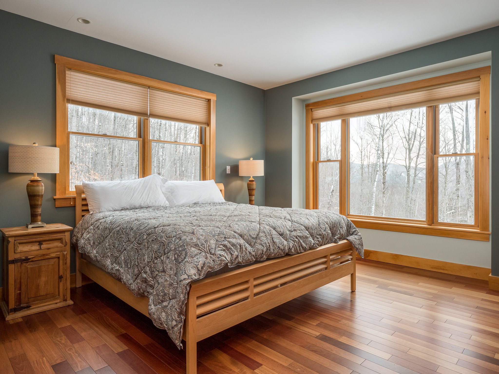 Bedroom with wooden bed, patterned comforter, two bedside tables with lamps, large windows with forest view, and hardwood floor.