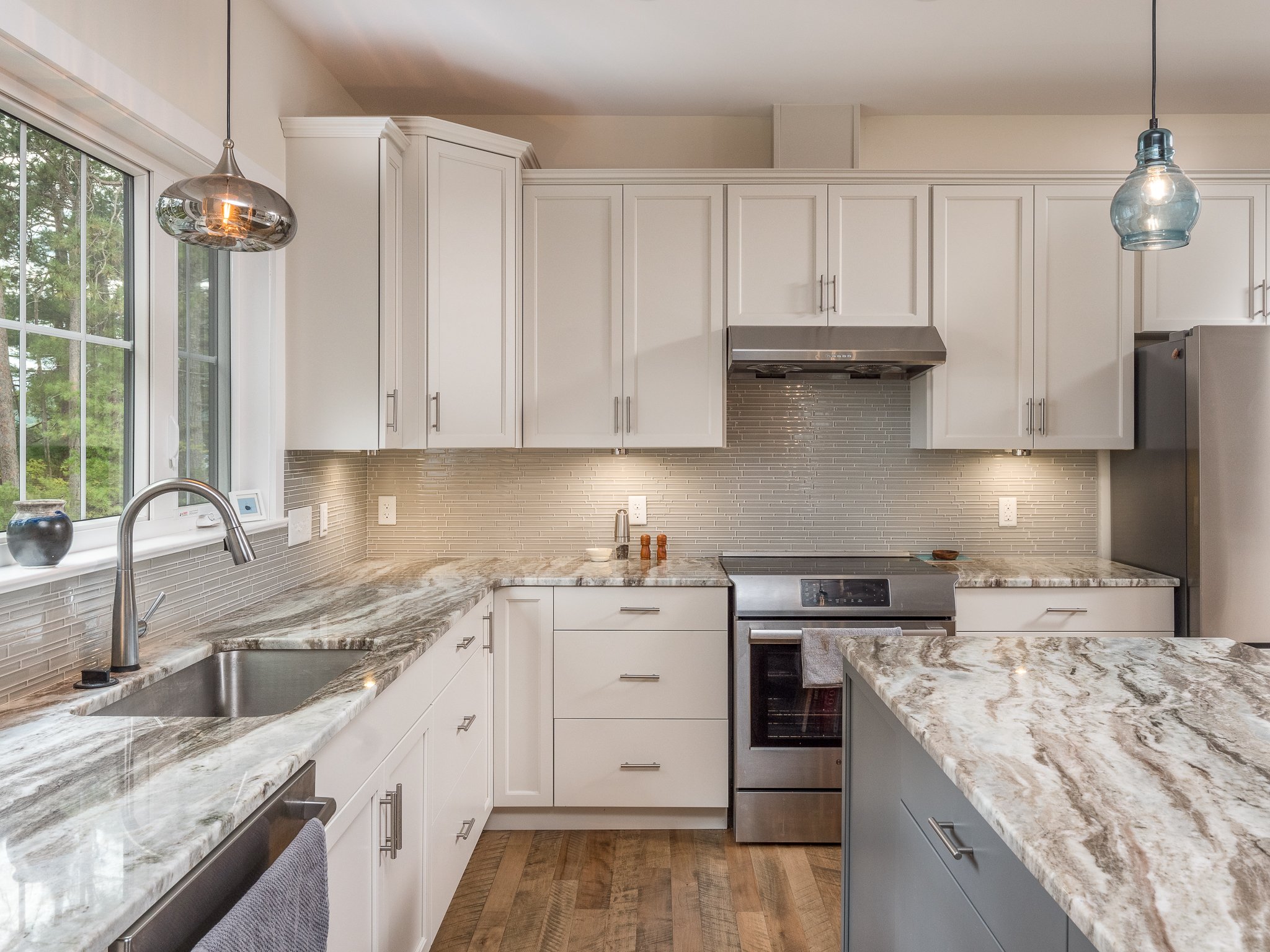 Modern kitchen with white cabinets, stainless steel appliances, under-cabinet lighting, granite countertops, pendant lights, and a window with outdoor view.