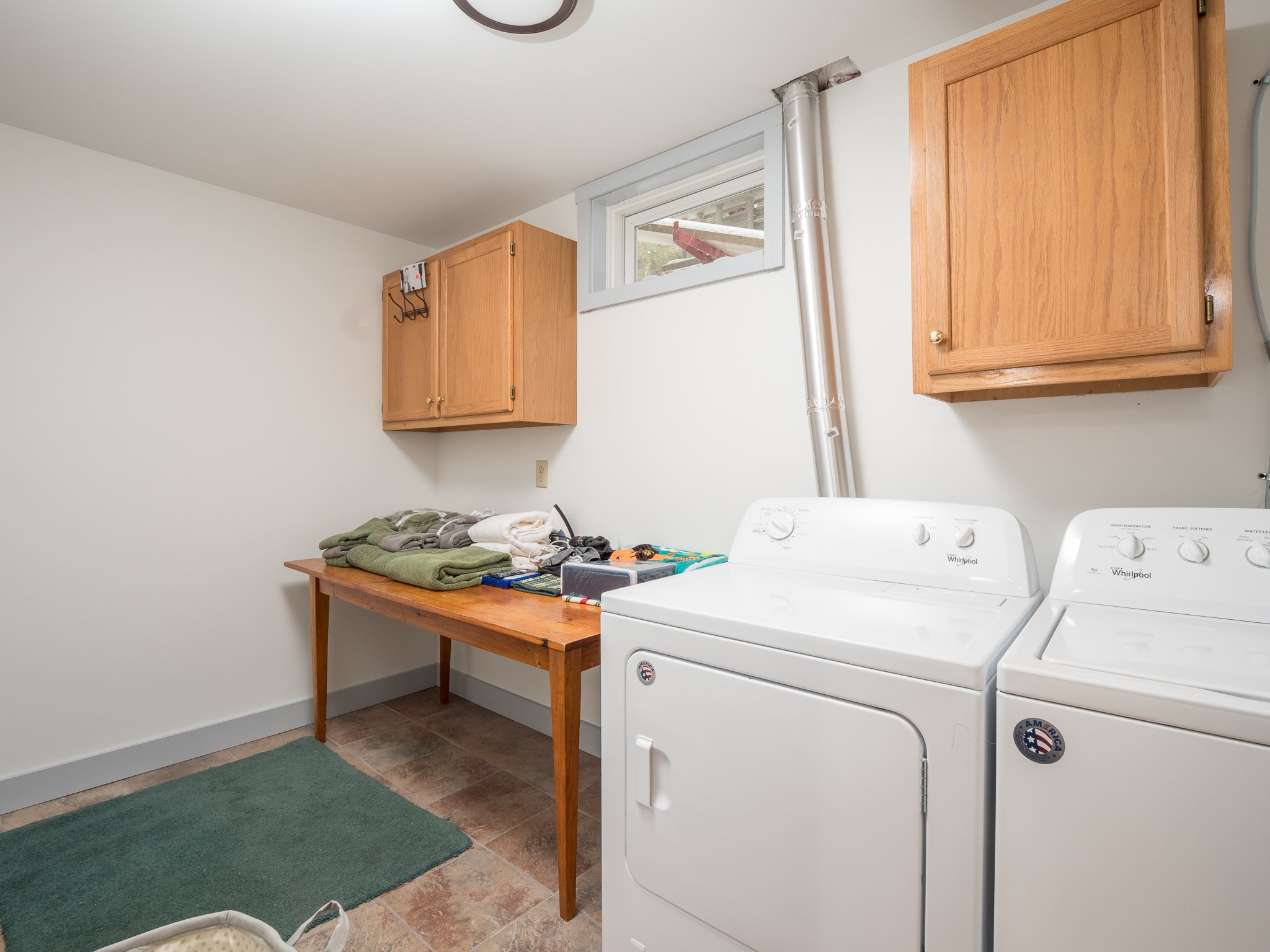 Laundry room with white washer and dryer, wooden cabinets, wooden table with folded clothes, and a green rug on tiled floor.