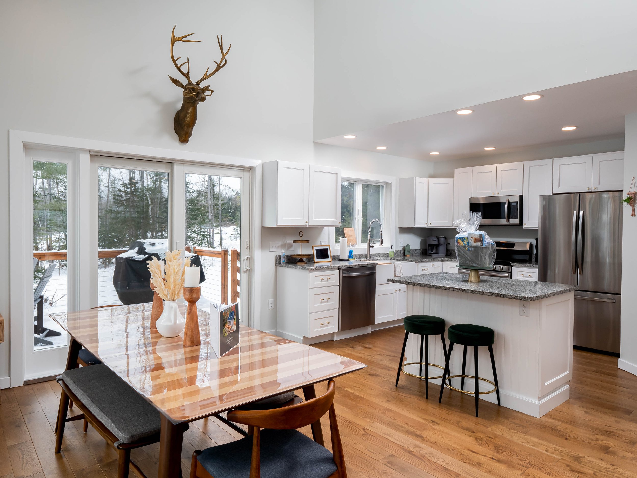 Modern kitchen with white cabinets, granite countertops, stainless steel appliances, and a wooden dining table with black and wooden chairs. A large sliding glass door leads to an outdoor deck with snow and trees visible outside. A mounted deer head