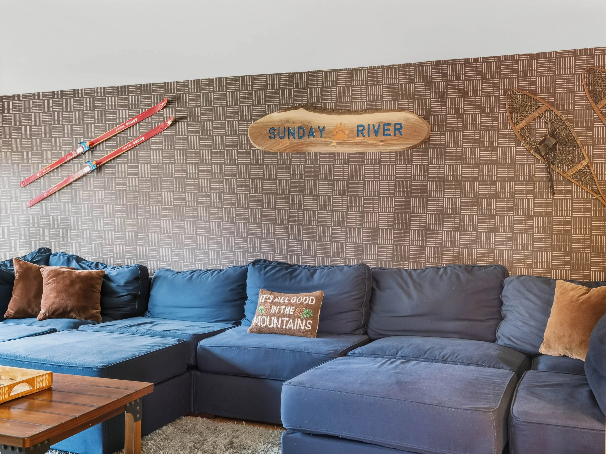 Living room with dark blue sectional sofa, brown and blue pillows, a wooden coffee table, and a decorative wall with ski and snowshoe decor, and a sign that says 'Sunday River'.