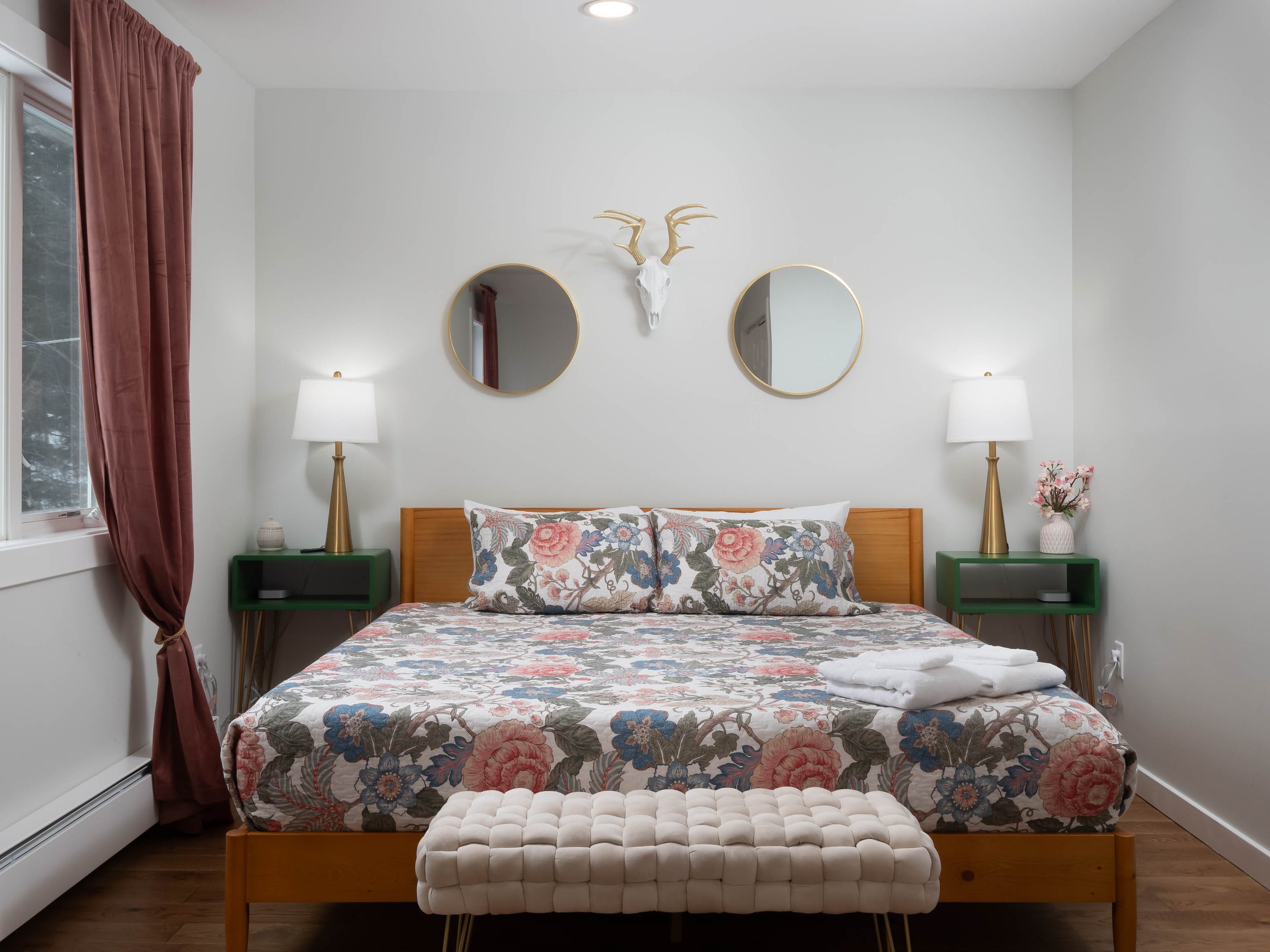 A bedroom with a wooden bed, floral bedding, two green nightstands with lamps, two round mirrors, a deer skull wall decor, a window with pink curtains, and a tufted bench at the foot of the bed.