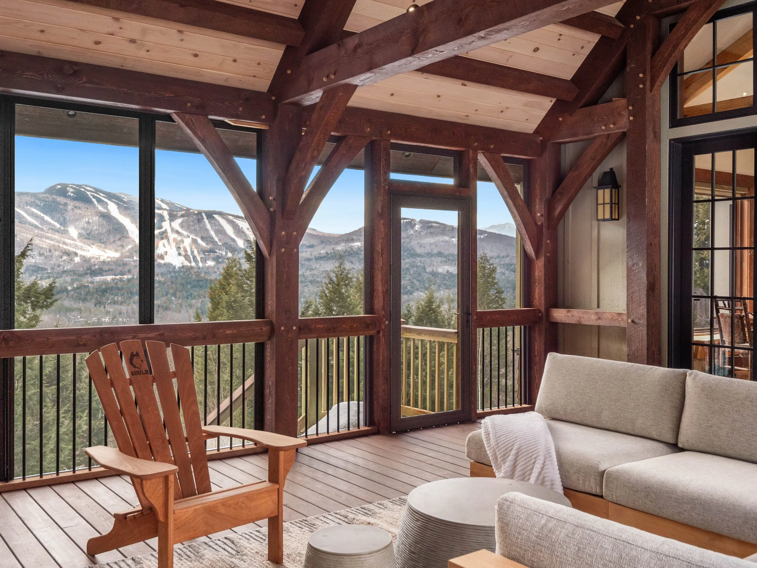 Living room with wooden beams, large windows showing snowy mountain views, beige couch with a white blanket, wooden Adirondack chair, and a small round coffee table.