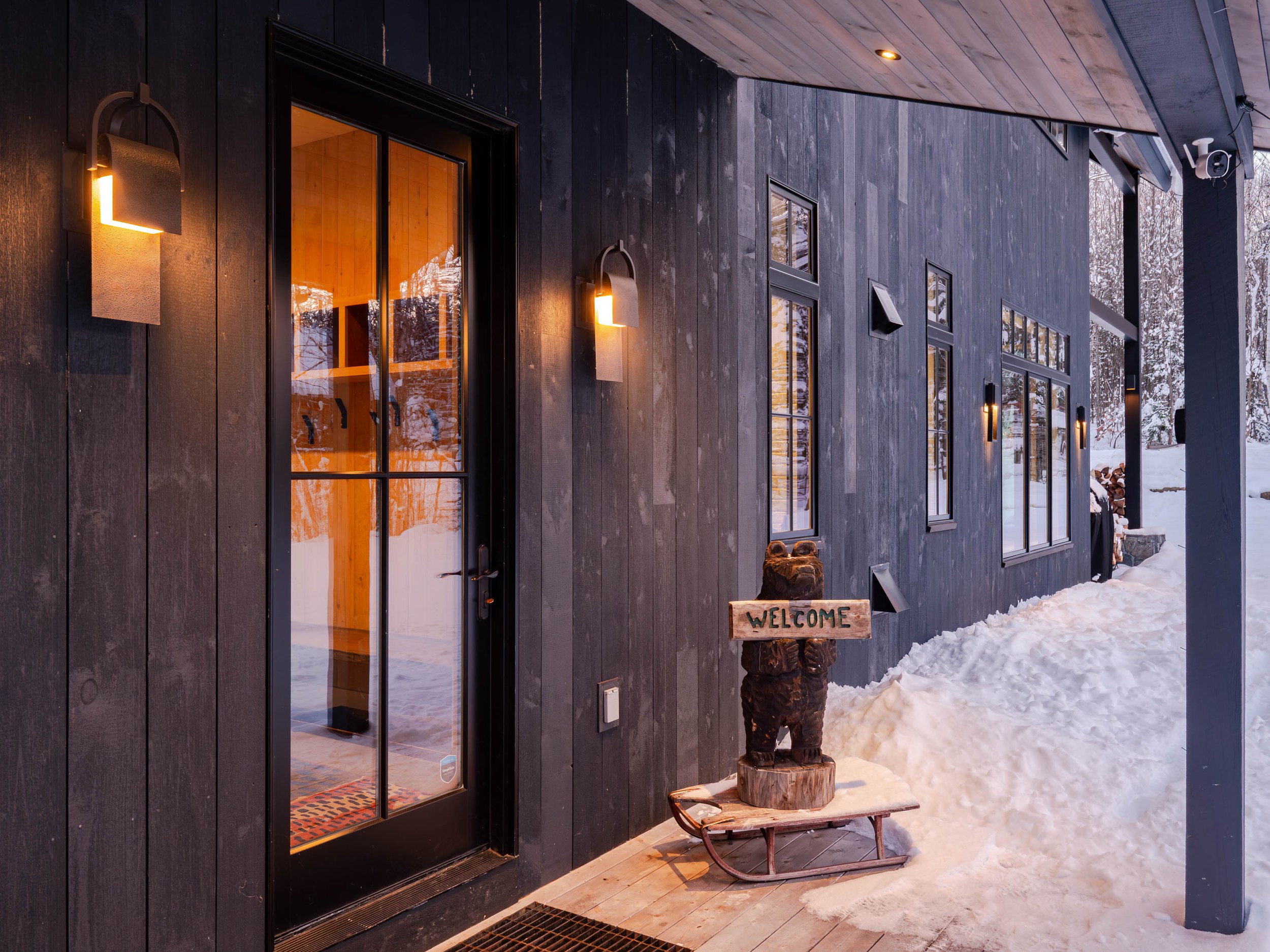 Modern cabin entrance with black siding, glass door, snow-covered surroundings, wall-mounted lights, and a bear sculpture with a welcome sign.