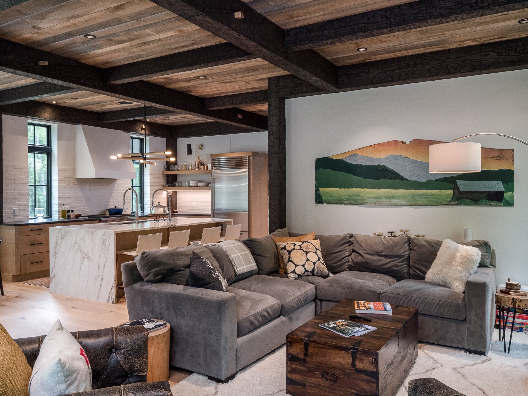 Modern living room with gray sectional sofa, wooden coffee table, artwork on wall, open kitchen with island, pendant lighting, and wooden ceiling beams.