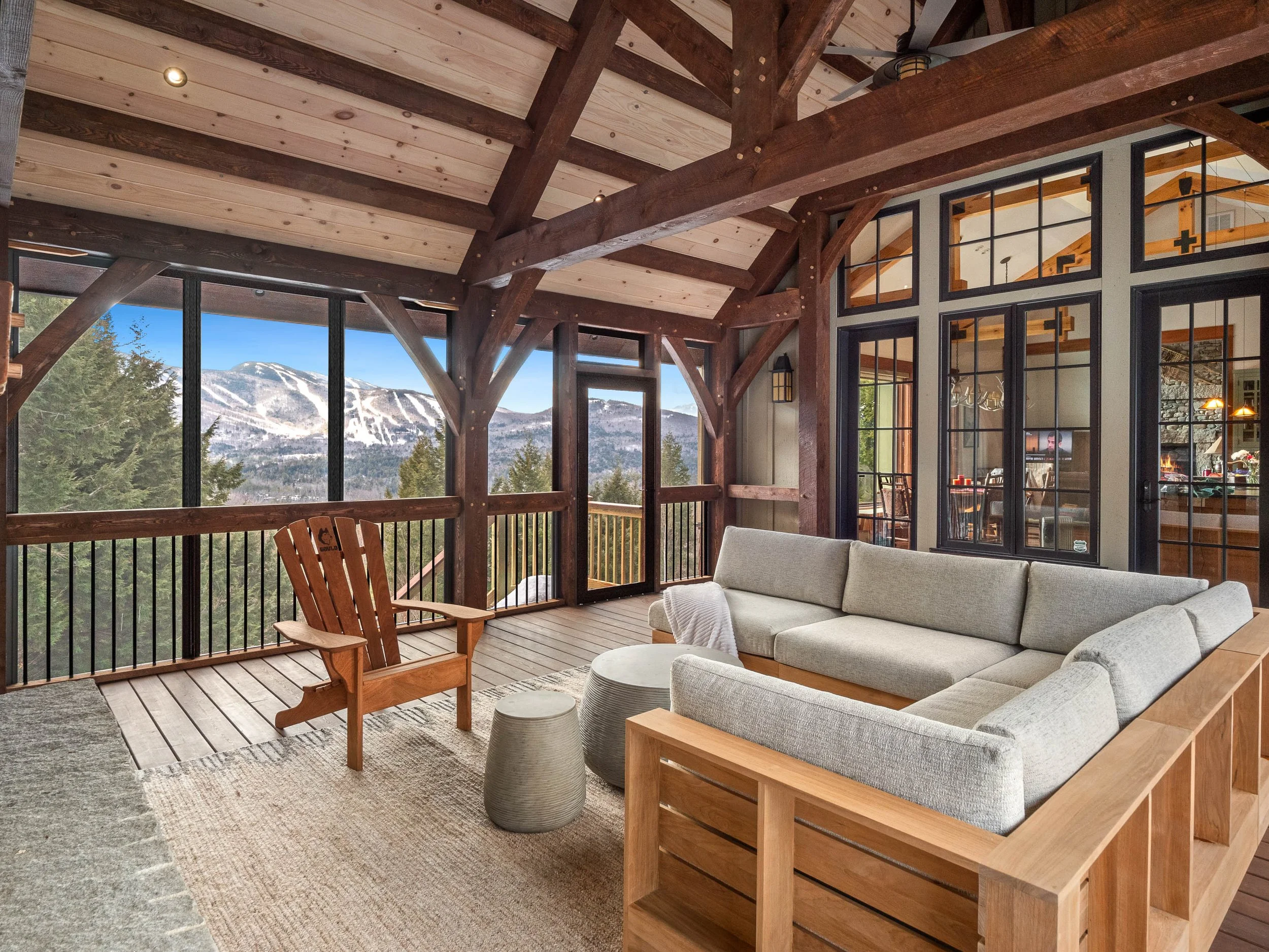 Living room with a screened balcony featuring wooden beams, a beige sectional sofa, a wooden Adirondack chair, small round tables, and large windows showing a mountain view.