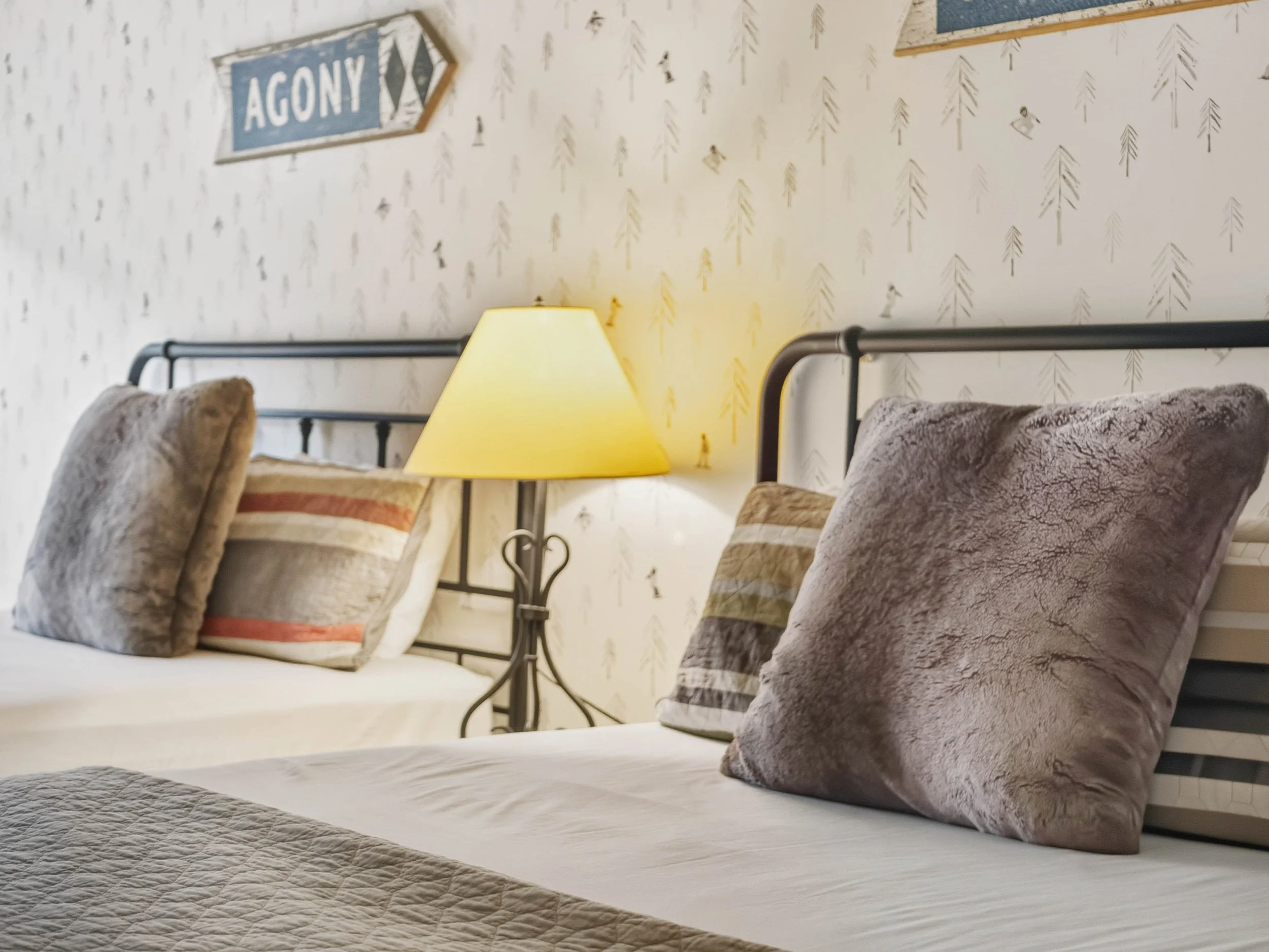 Two beds with plush pillows and beige bedding, a bedside lamp with a yellow shade, and a wall with a nature-themed wallpaper and a sign that says 'AGONY' with arrows.