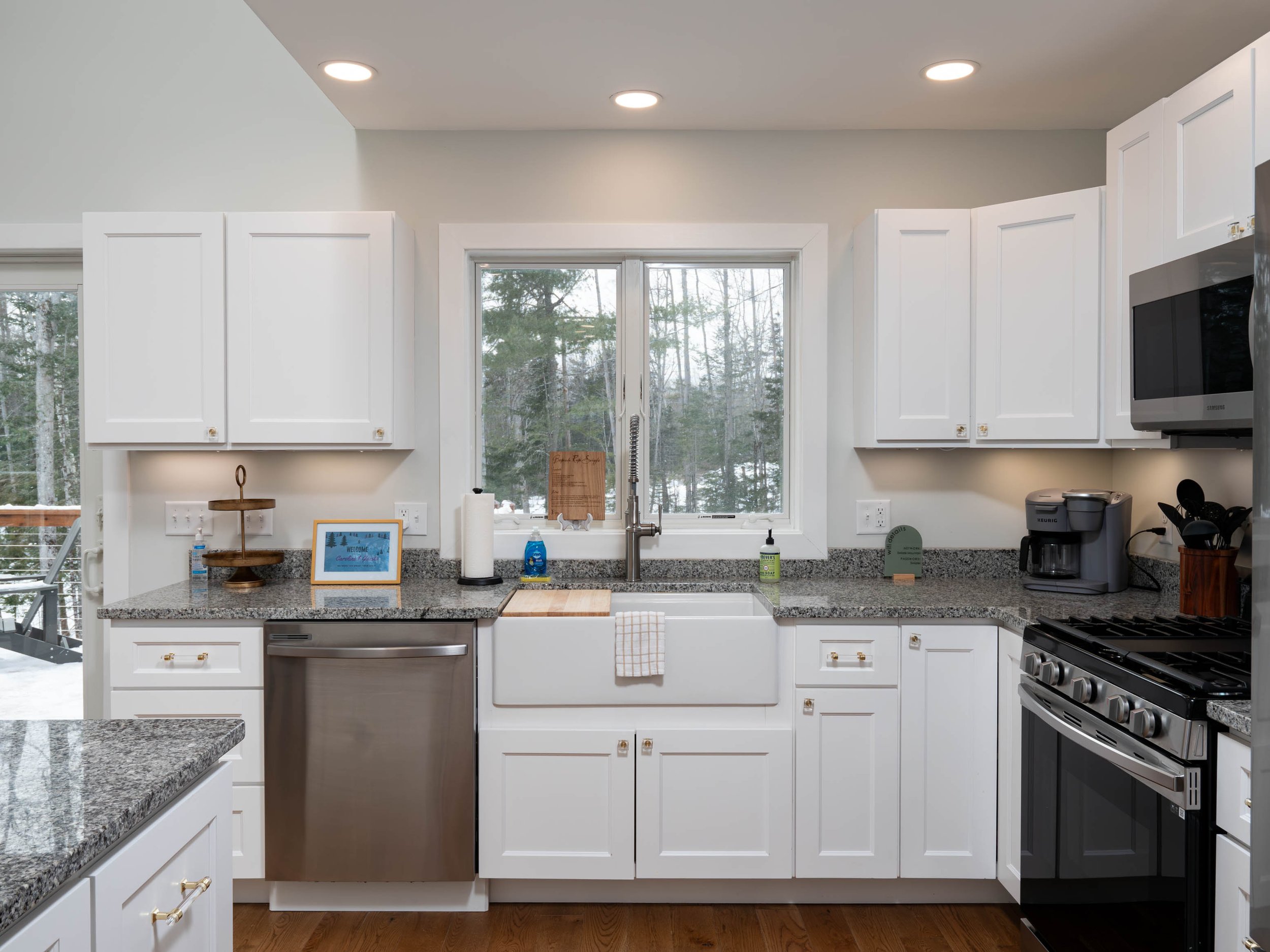 Kitchen with white cabinets, gray granite countertops, a large window above the sink, stainless steel dishwasher, black stove, microwave, coffee maker, and a view of trees outside.