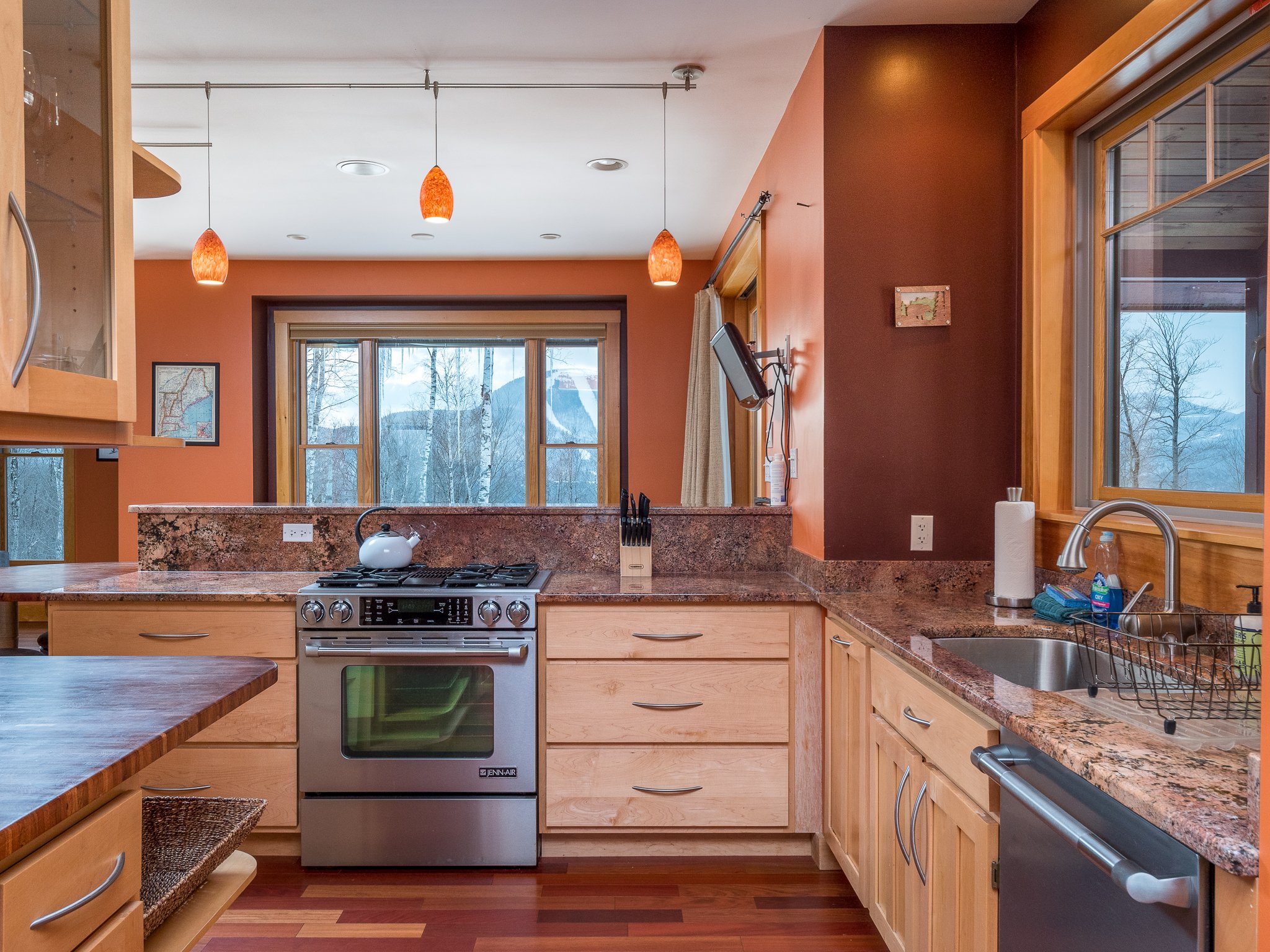 Modern kitchen with wooden cabinets, granite countertops, stainless steel stove, and sink. Large window with winter landscape view, orange pendant lights, and hardwood flooring.
