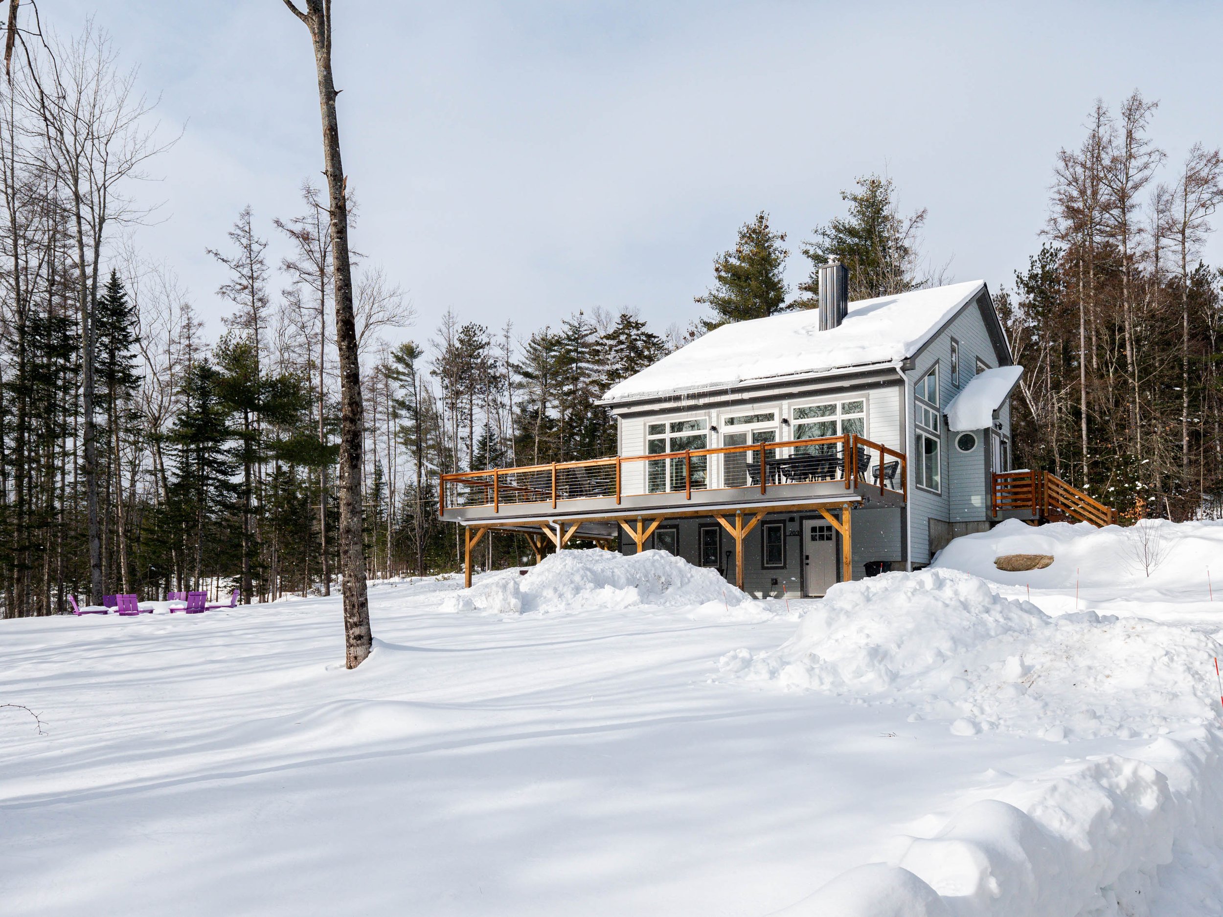 A house in a snow-covered landscape with a large deck, surrounded by trees, in winter.