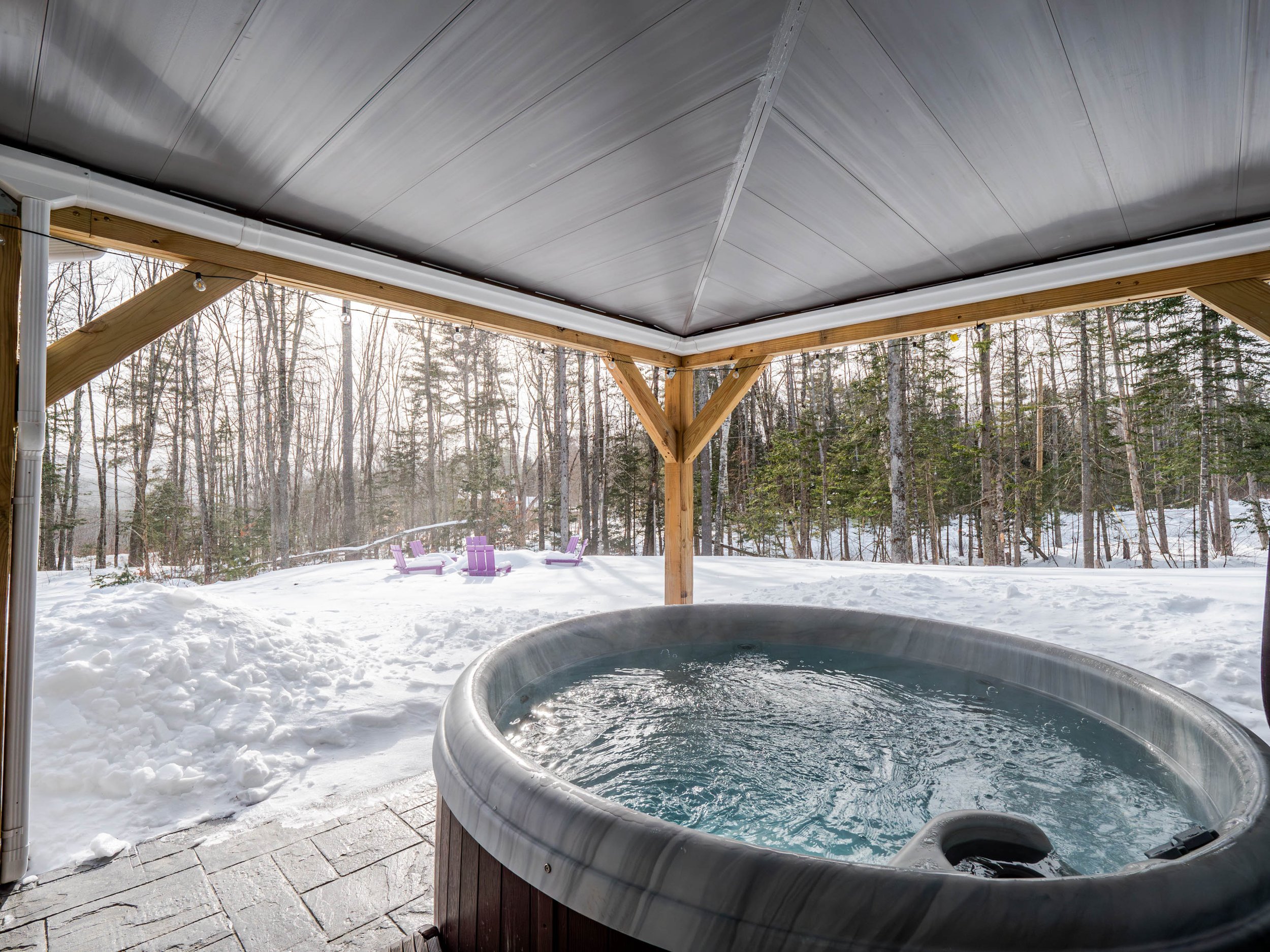 Outdoor hot tub on a covered patio, overlooking a snowy wooded yard with purple Adirondack chairs in the background.