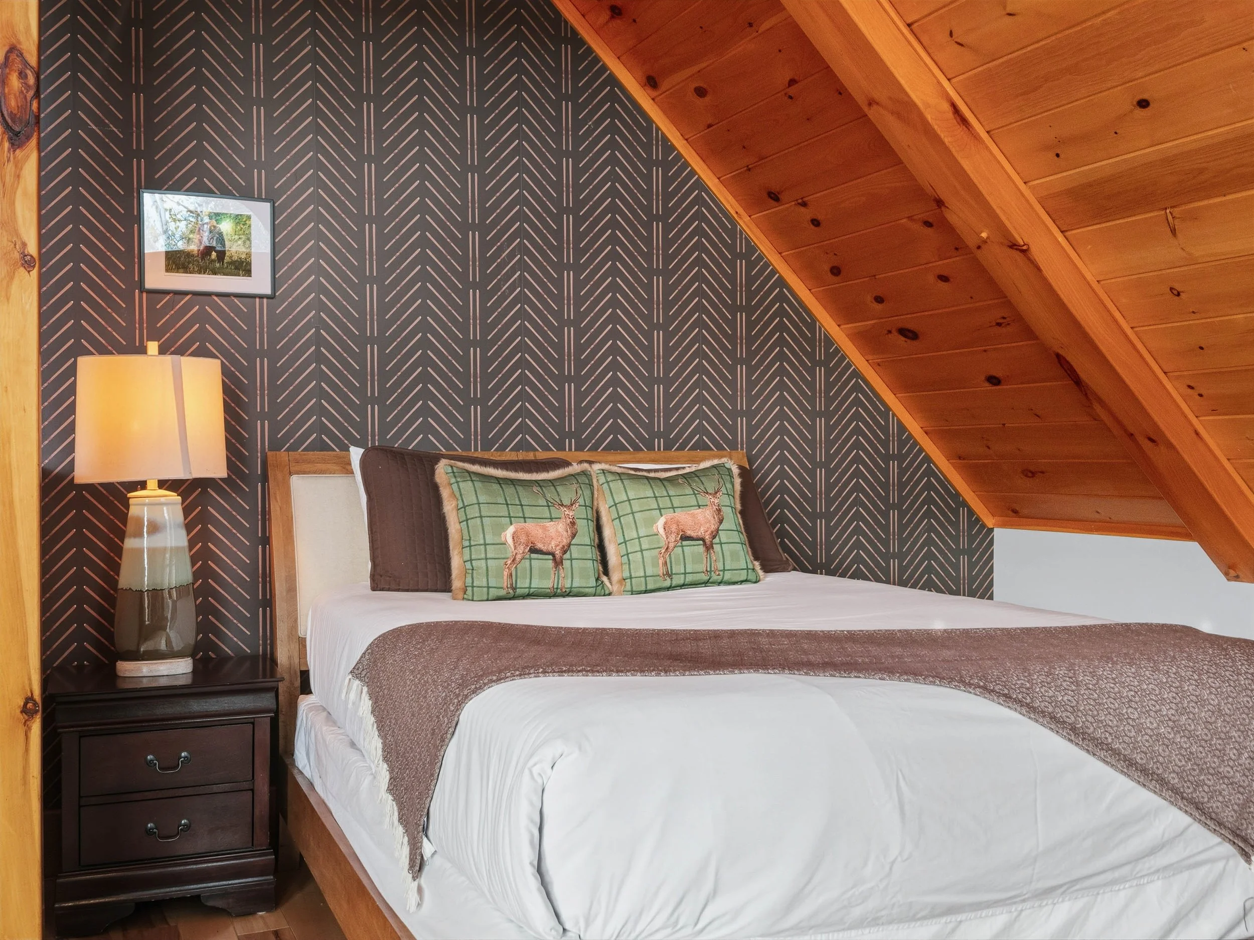 A cozy bedroom in a rustic cabin with a sloped wood-paneled ceiling, a bed with deer-themed pillows, a dark nightstand with a lamp, and a framed picture on the wall.
