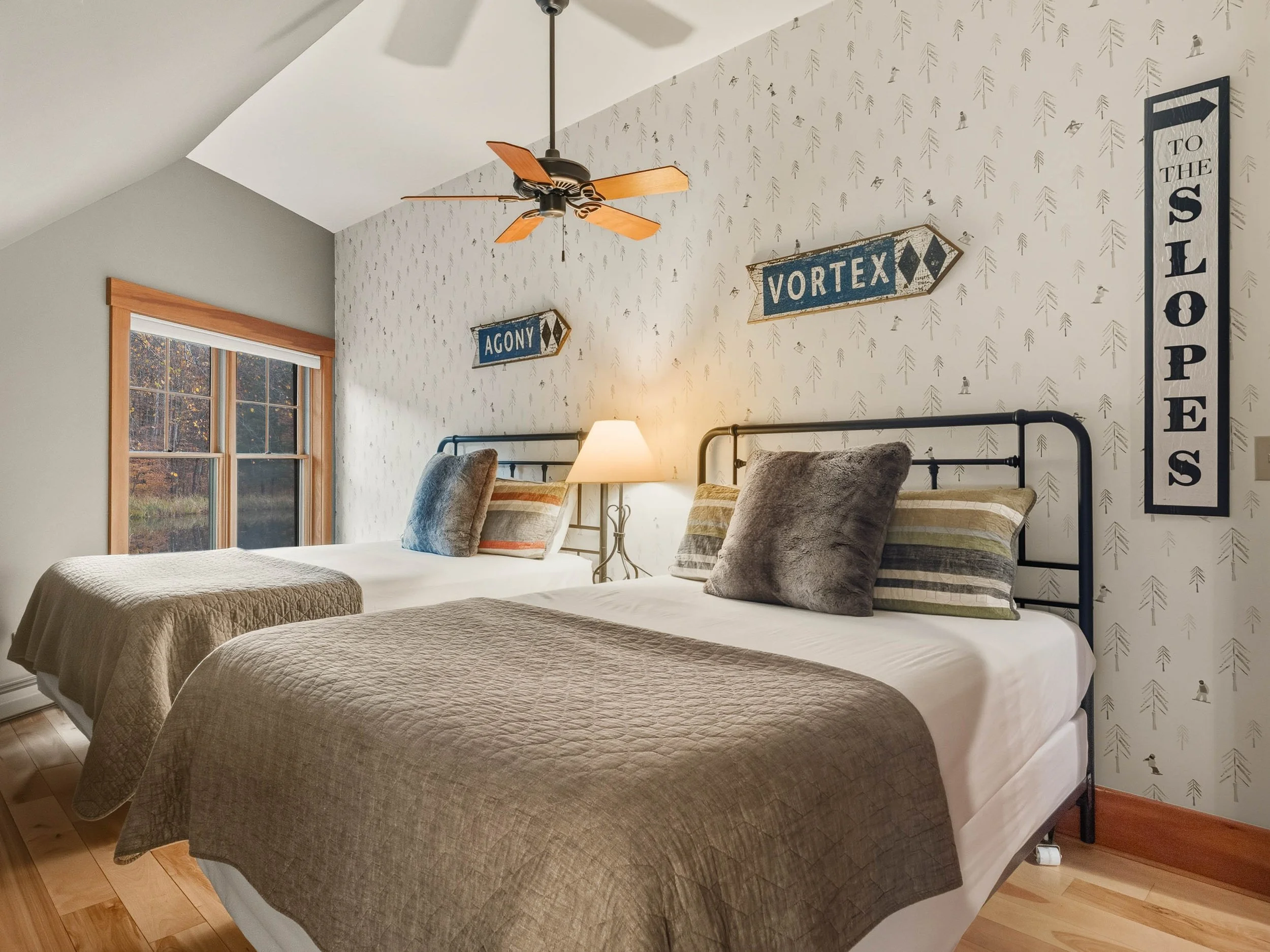 Two beds with dark metal headboards in a bedroom with woodland themed wallpaper. One bed has beige and brown bedding, the other has cream and striped bedding. Decorative pillows on each bed. A window with natural wood trim and a ceiling fan are in th