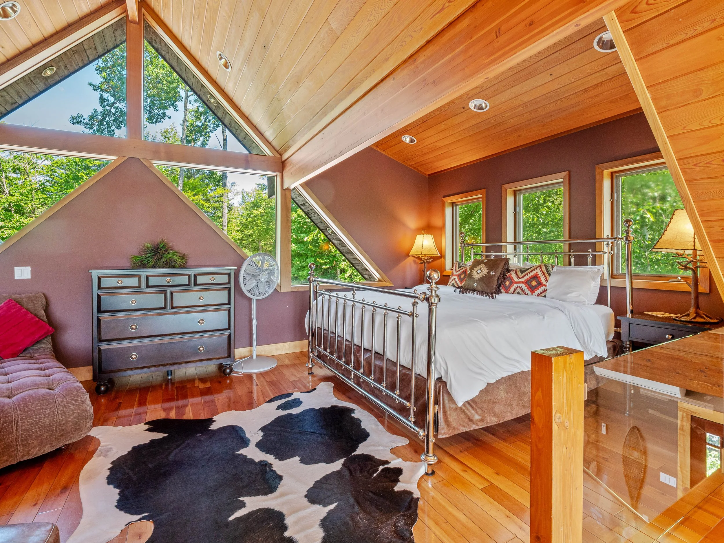 Bedroom with large windows showing green trees, a metal bed frame with white bedding, wooden floors, a cowhide rug, a dresser, and lamps with warm lighting.