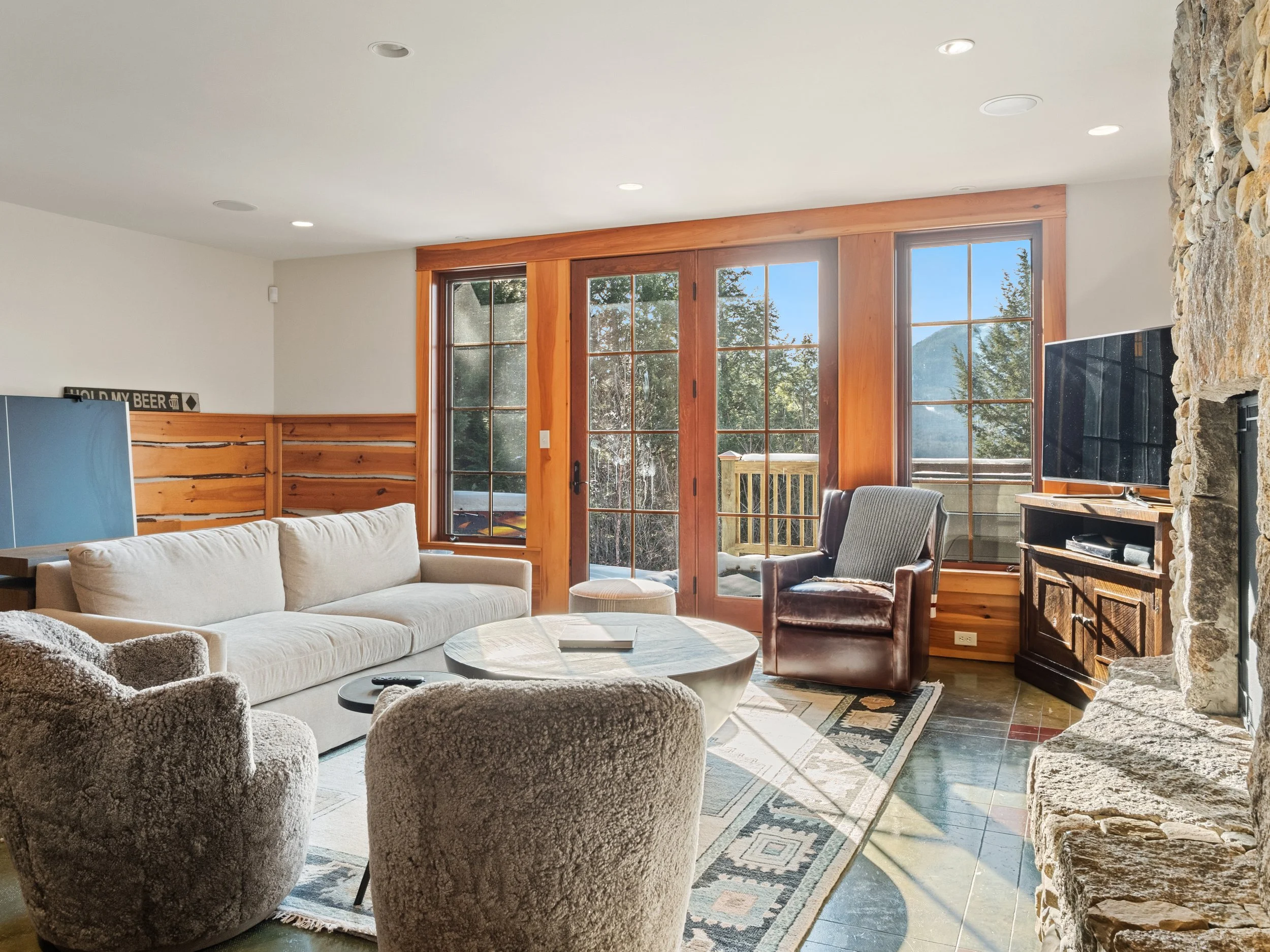 Cozy living room with large windows, stone fireplace, cream sofa, plush armchairs, and a leather armchair, with mountain view outside.
