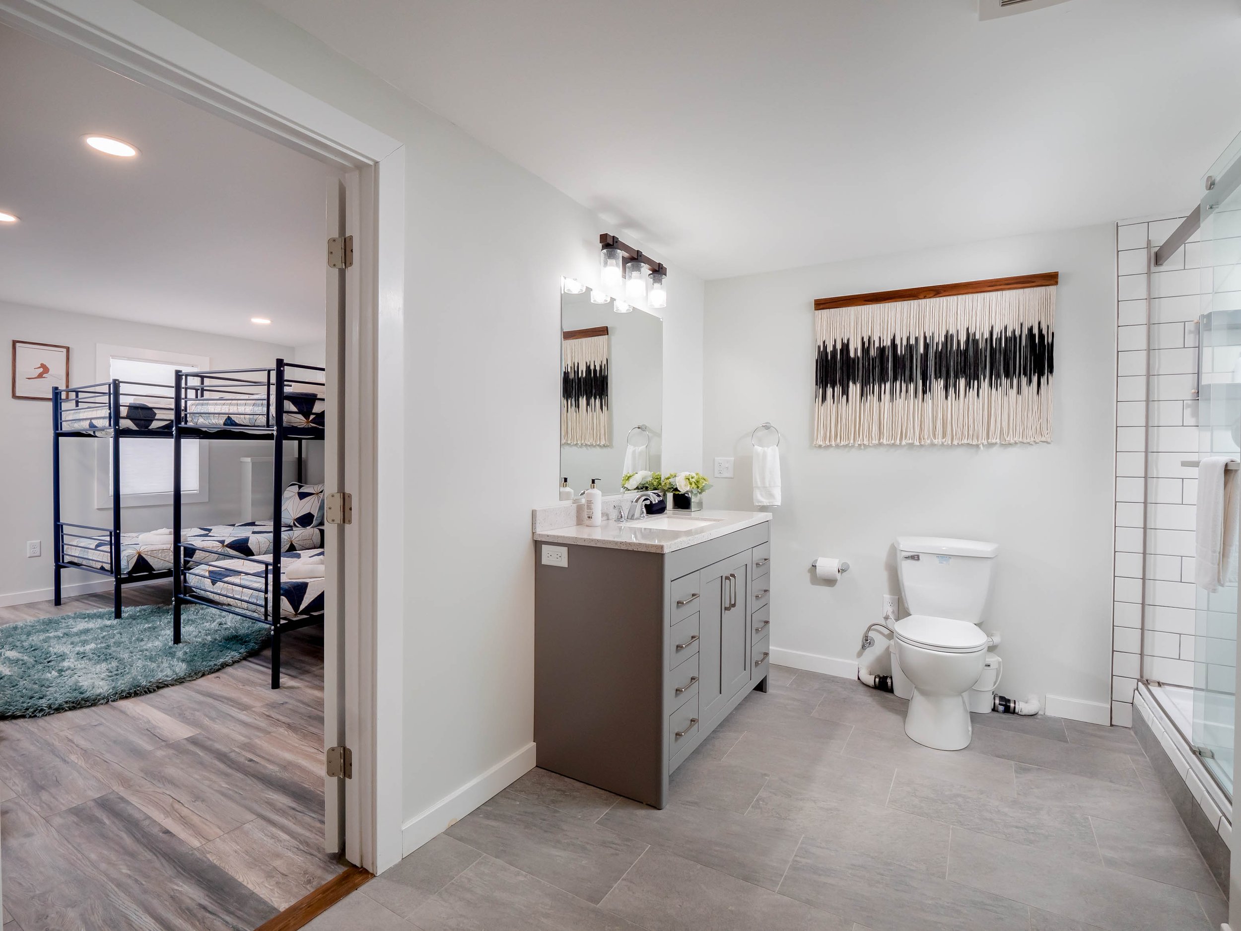 Bathroom with gray vanity, large mirror, white walls, black and white wall hanging, toilet, towel rack, and walk-in shower with glass enclosure. Adjacent bedroom with bunk beds and framed artwork.