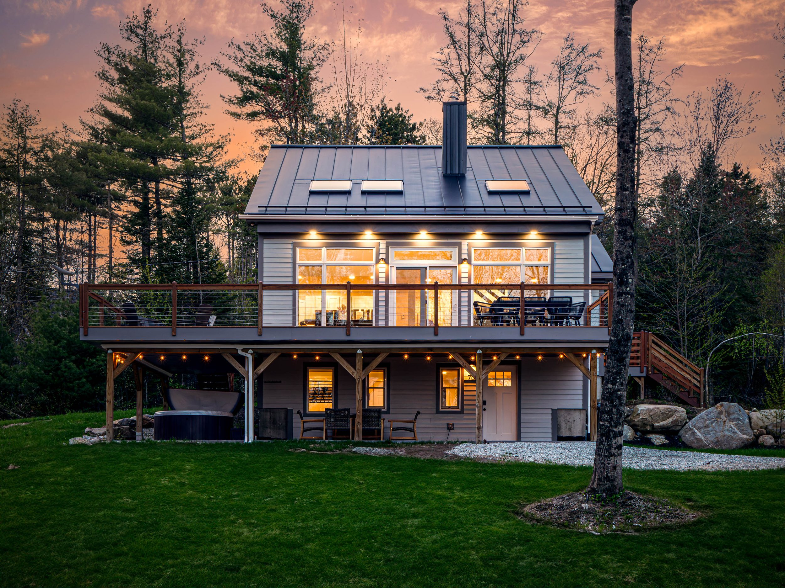 A two-story house with a metal roof, exterior lights on, and large windows illuminated from inside, situated in a wooded area during sunset. The house features a balcony and outdoor seating with a hot tub underneath.