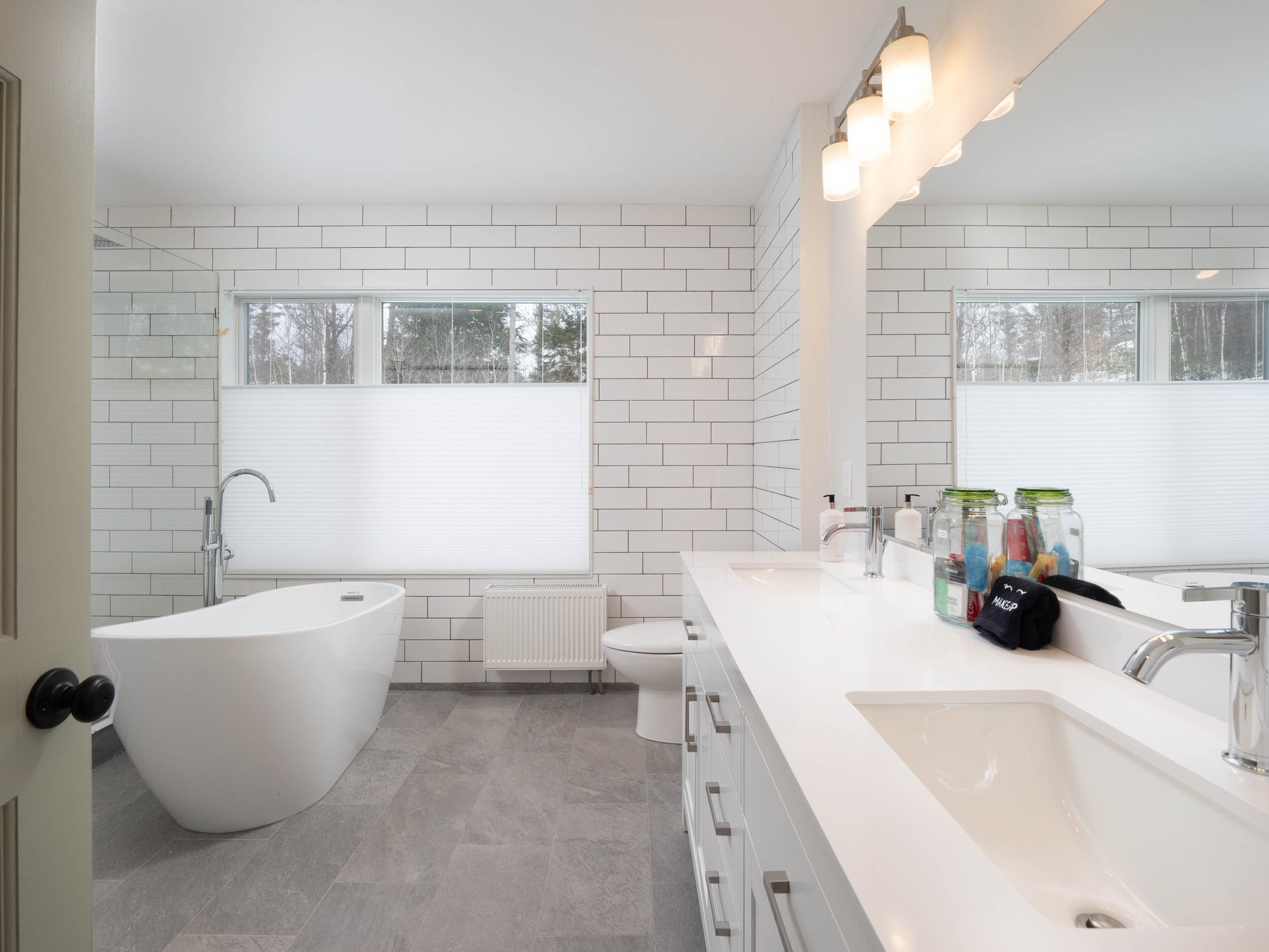 Swizzle Mountain Lodge's large modern bathroom with white tiled walls, a freestanding bathtub, large mirror, double vanity, and gray tiled floor.