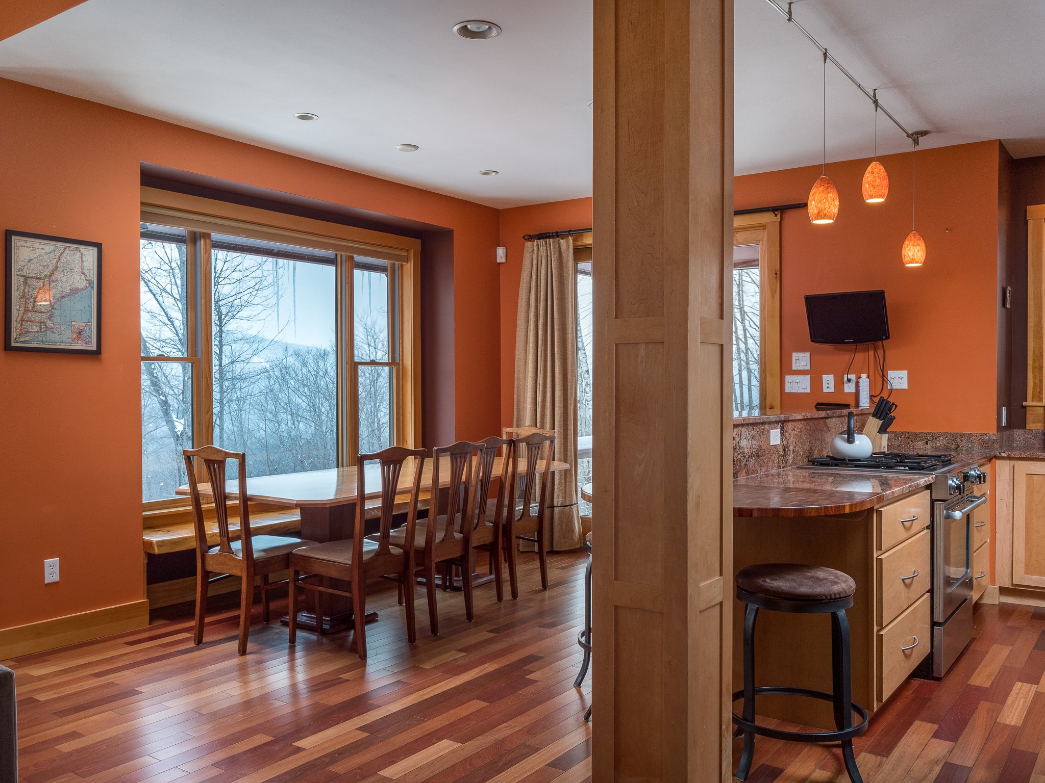 Modern kitchen and dining area with orange walls, wooden flooring, large windows, a rectangular dining table with chairs, pendant lights, and kitchen countertops.