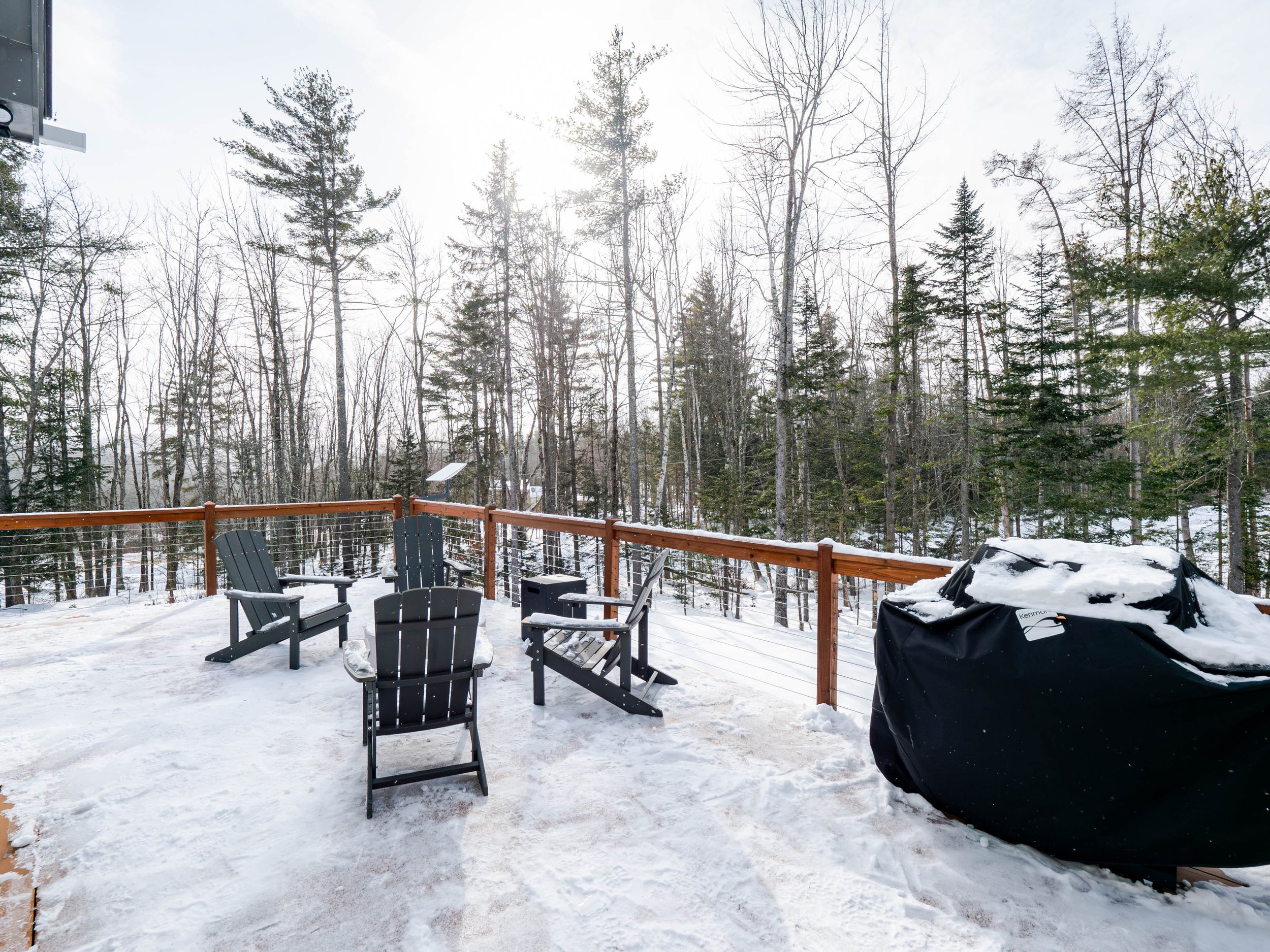 Snow-covered outdoor deck with black Adirondack chairs and a covered grill, surrounded by a wooden railing, overlooking a forest with leafless and evergreen trees under a cloudy sky.