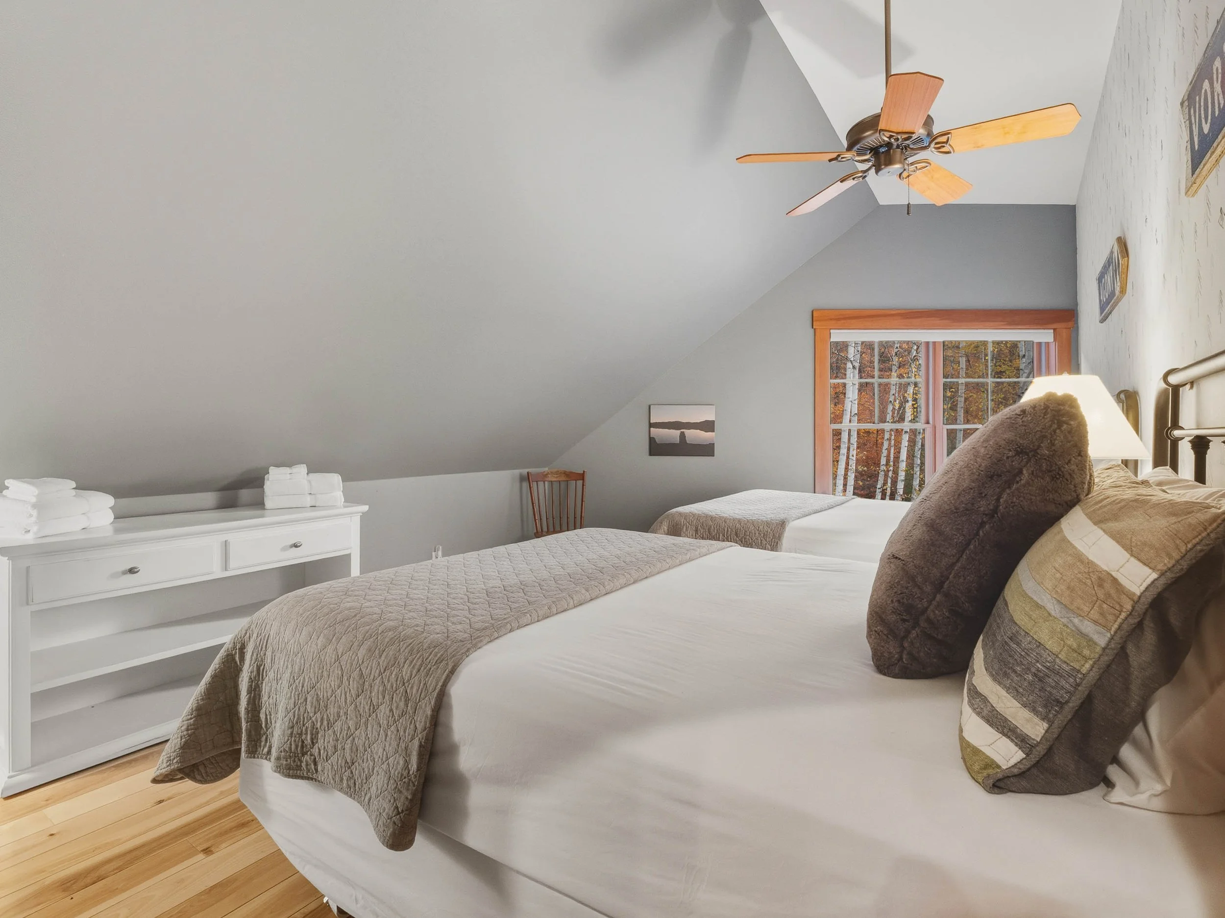 A cozy attic bedroom with two beds, a ceiling fan, a window looking out to trees, pillows, a lamp, and a nightstand.