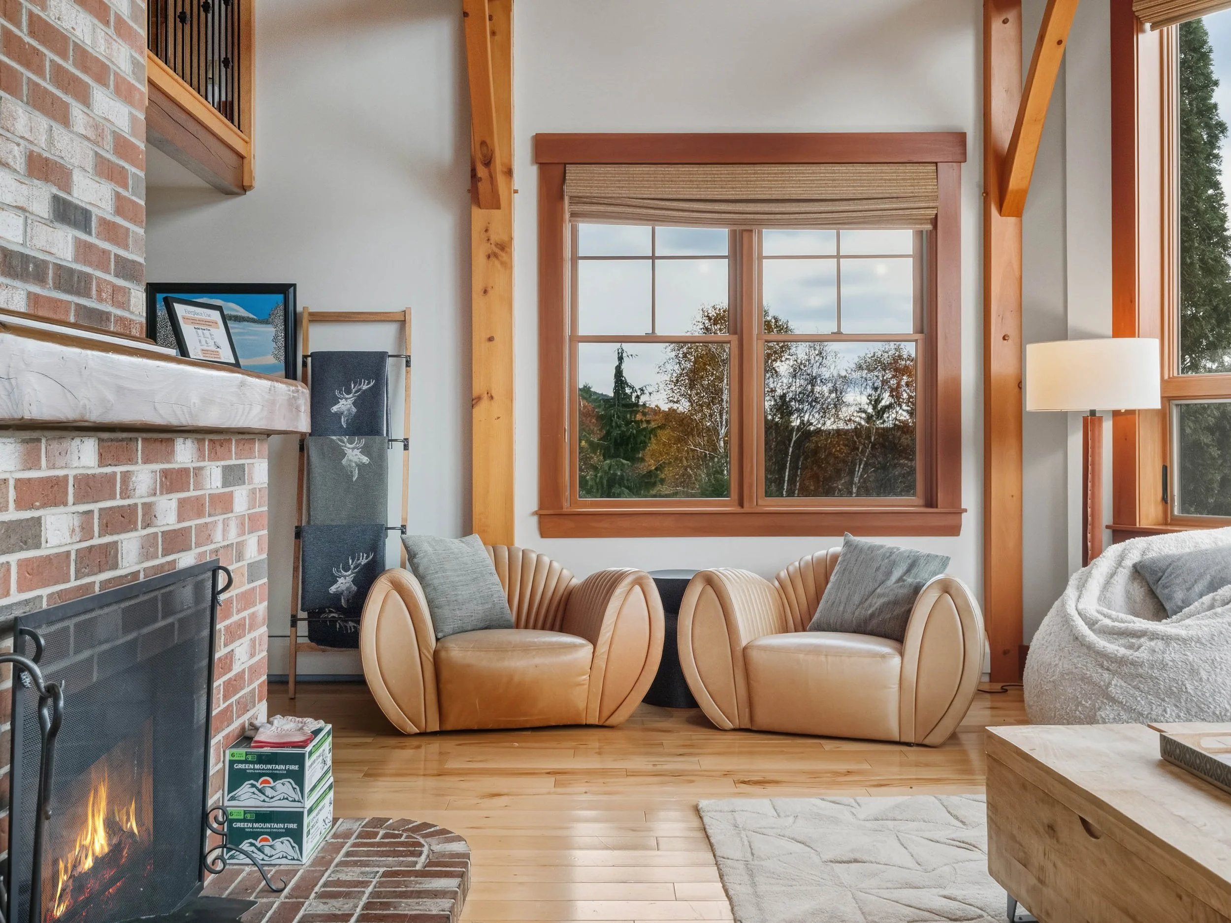 Living room with wood floors, brick fireplace, two tan leather lounge chairs with cushions, a bean bag chair, large windows showing trees outside, a floor lamp, and a wooden coffee table.