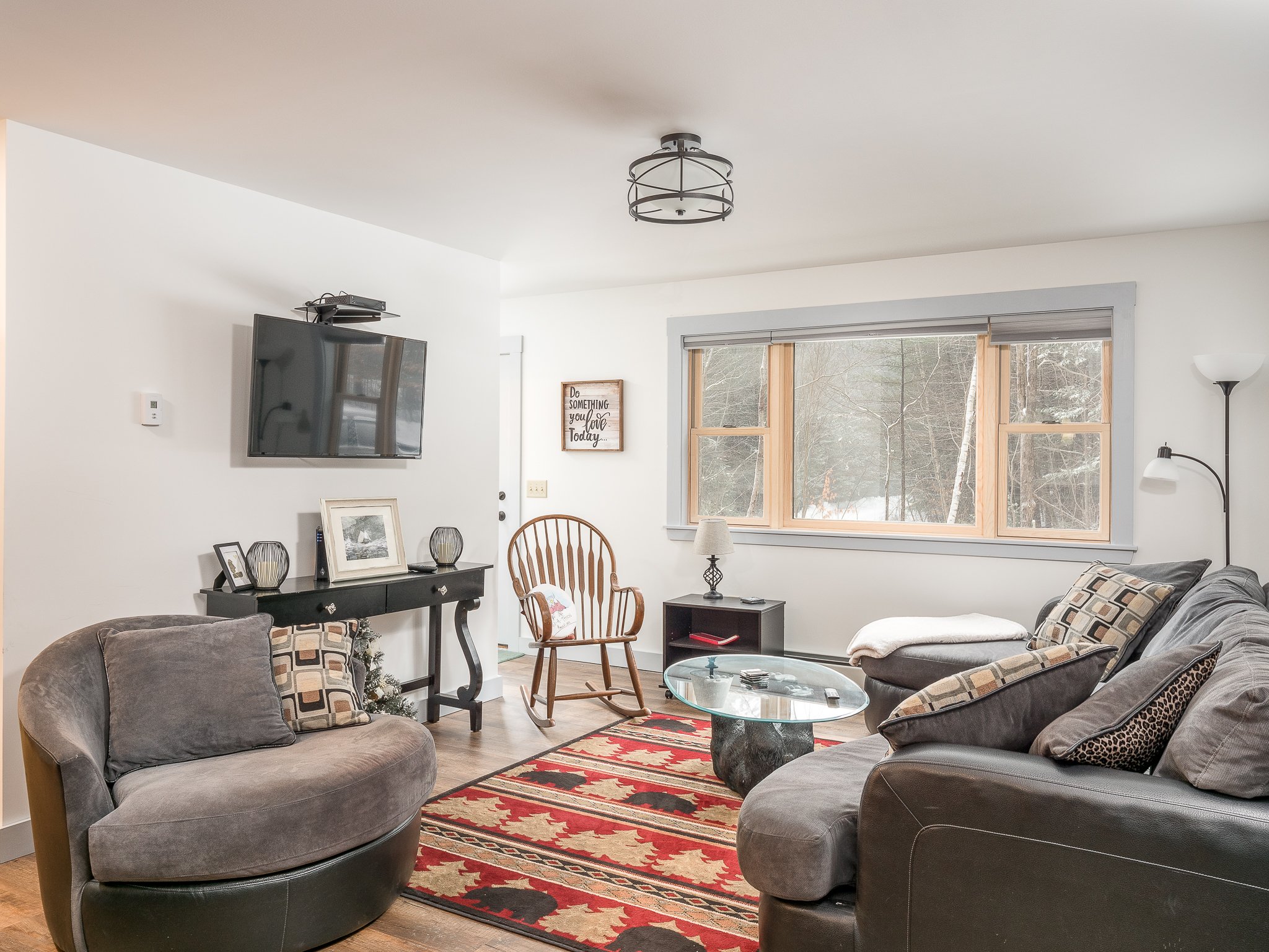 Cozy living room with modern decor, featuring a wall-mounted flat-screen TV, a plush sofa, and an armchair. The room includes a coffee table, patterned rug, large window with scenic view, and decorative items on a console table.