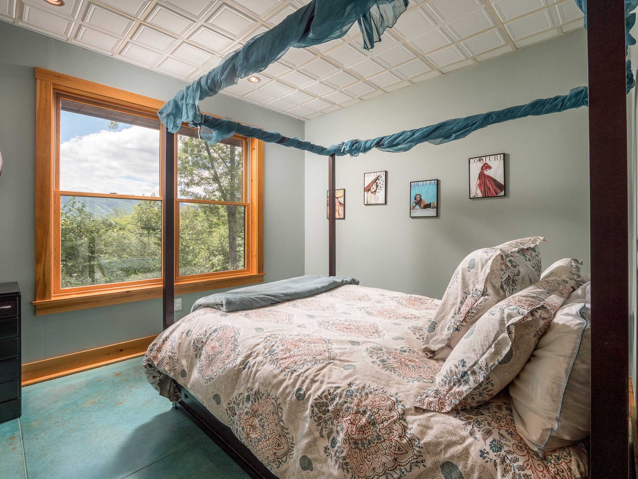 Bedroom with a four-poster bed, patterned bedding, teal fabric canopy, large window with scenic view, and fashion-themed wall art.